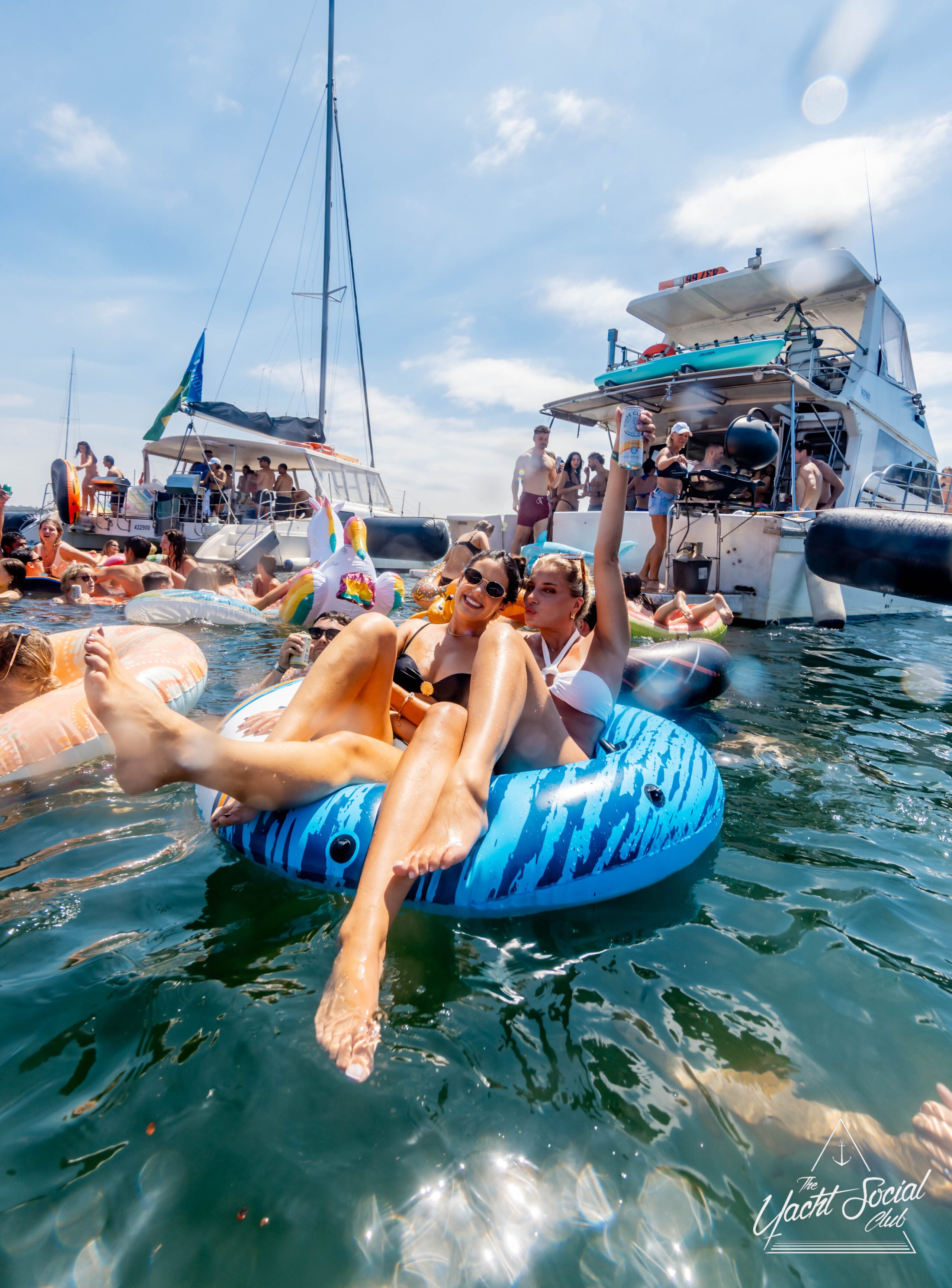 A group of people enjoying a sunny day on the water. Two individuals sit on a blue inflatable, surrounded by others on floaties and boats. The background features yachts and a bright blue sky. The atmosphere is lively and festive.