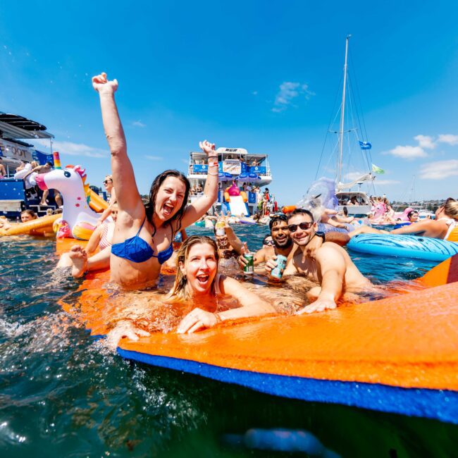 A group of people laughing and enjoying themselves in the water, surrounded by inflatable pool floats, including a unicorn. They appear to be celebrating, with one person raising their arm in excitement. The sky is clear and sunny.