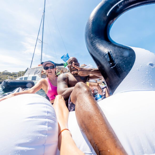 Two people are enjoying a sunny day on the water, lounging on a large inflatable swan float. A sailboat is visible in the background, with clear blue skies and a shoreline in the distance.