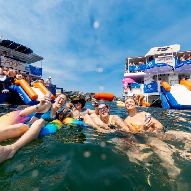 A group of people relax in the water on inflatable floats, smiling and holding drinks. Behind them, two yachts are docked with more people enjoying the sunny day. Some guests are on slides and others are seated on the yachts.