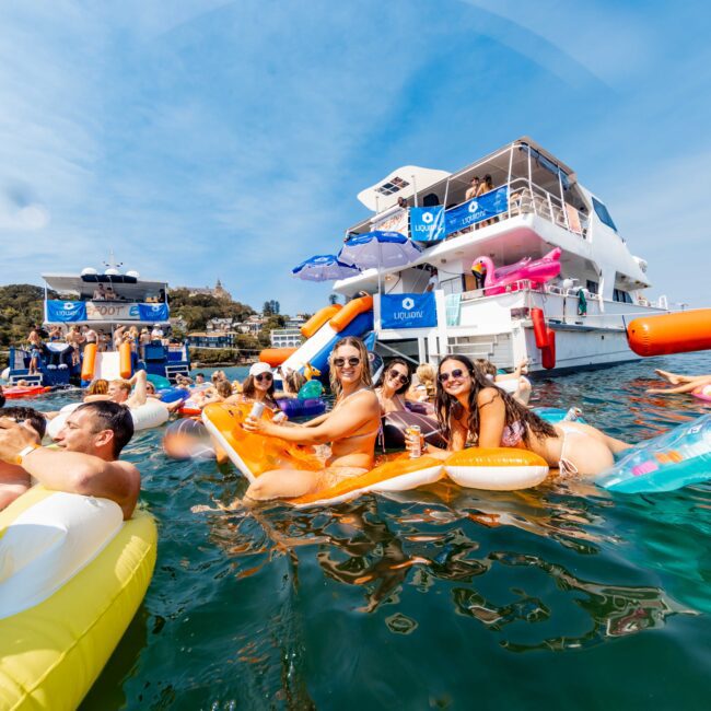 People relaxing on inflatable tubes in the water near two yachts on a sunny day. The group is smiling and enjoying themselves, with the yachts displaying festive banners. Hills and trees are visible in the background.