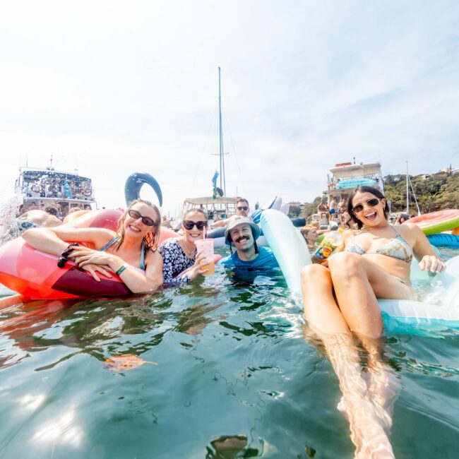 Group of people having fun in the water with inflatable floats near boats. They are smiling and relaxed, wearing swimwear under a clear blue sky. Hills and trees are visible in the background.