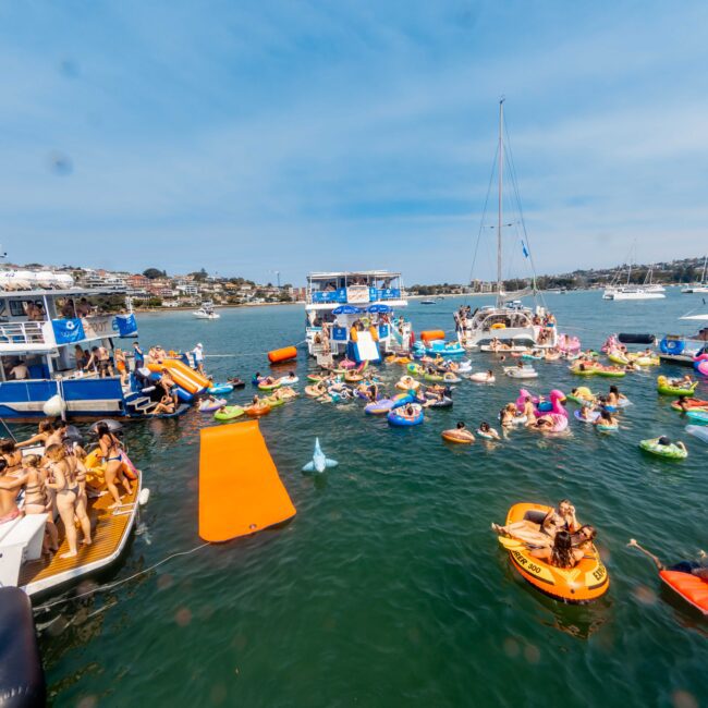 A lively scene on the water with numerous people on colorful inflatable rafts and boats. Many are wearing swimsuits and enjoying the sunny day. Surrounding them are several anchored boats. The sky is clear, and a coastal landscape is visible in the background.