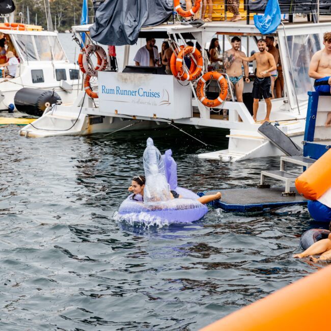 People on a boat party; some are wearing swimsuits and enjoying drinks. A person is riding an inflatable unicorn in the water, with splashing motion. Lifebuoys and a sign reading "Rum Runner Cruises" are visible on the boat. City skyline in background.