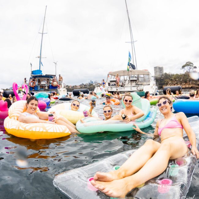 A group of people relax on colorful inflatable pool floats in the water, holding drinks and smiling. Boats and more people are visible in the background. The sky is overcast.