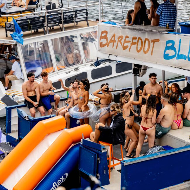 A group of people enjoying a sunny day on a boat named "Barefoot Blue". Some are sitting and chatting while others stand and dance. An inflatable slide is attached to the boat, and the water in the background is calm.