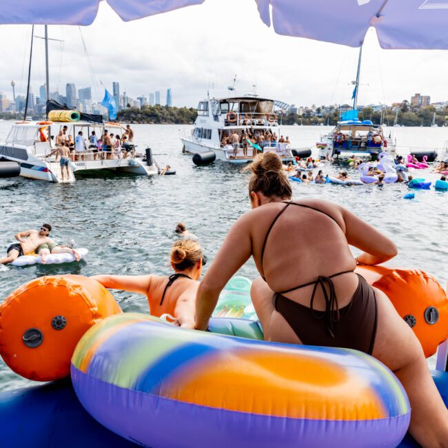 People enjoy a sunny day on the water, swimming and lounging on colorful inflatables near several boats. The city skyline is visible in the background, and a person holds a water bottle on a boat's deck.