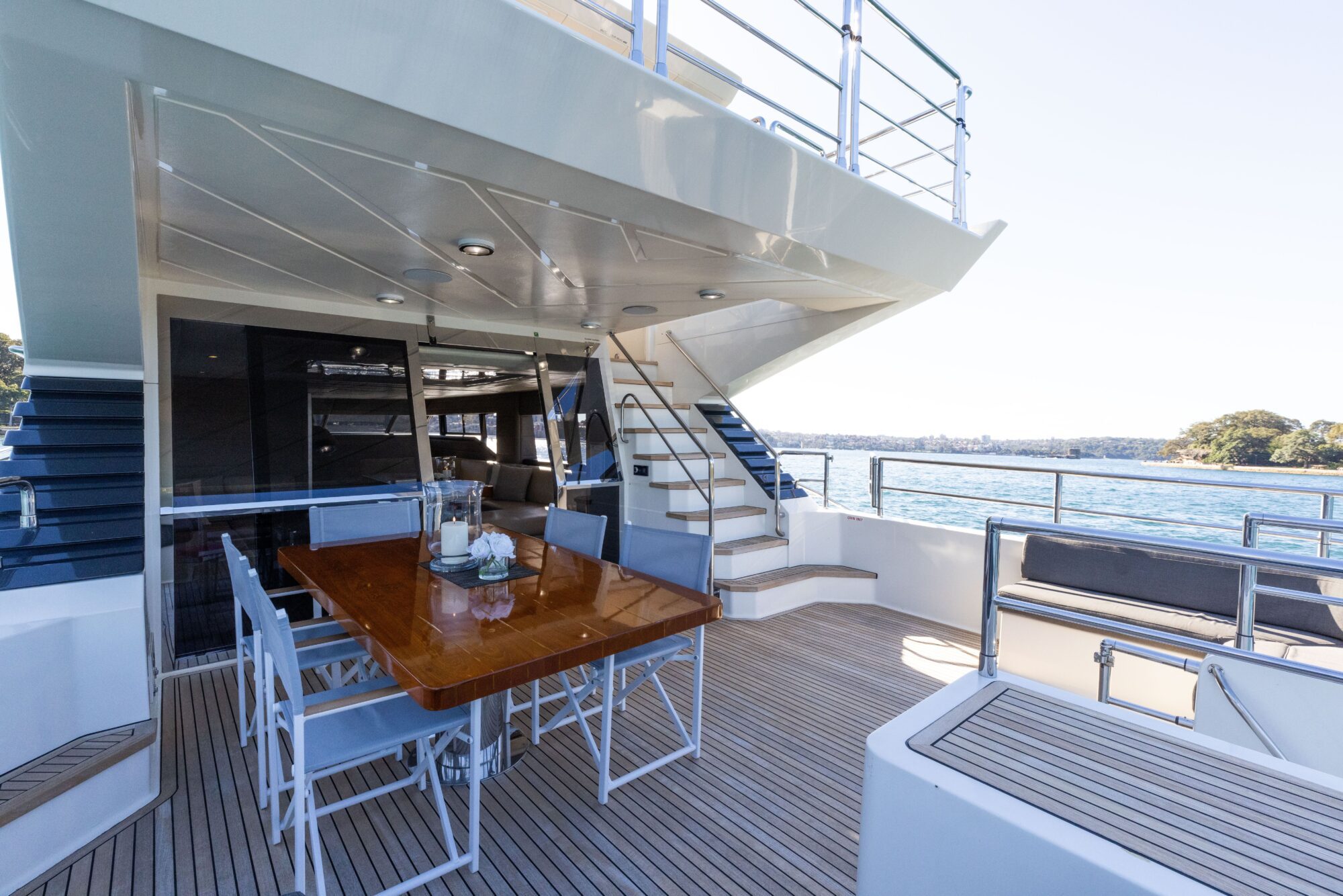 Outdoor dining area on a yacht featuring a polished wooden table with chairs, under a covered deck. Stairs lead to an upper level. The yacht is anchored near calm blue water with a distant shoreline.