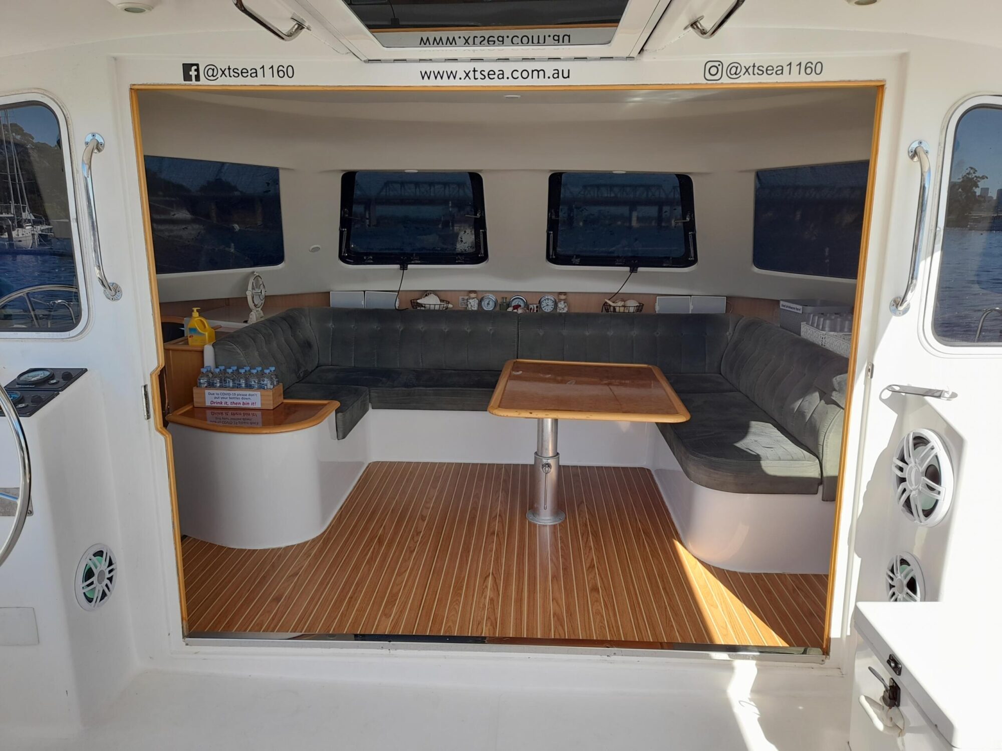 The image shows the interior of a boat with a cozy seating area, a wooden table, and cushioned benches lining the walls. Bottled water and snacks are arranged on a side counter, and large windows offer views outside.