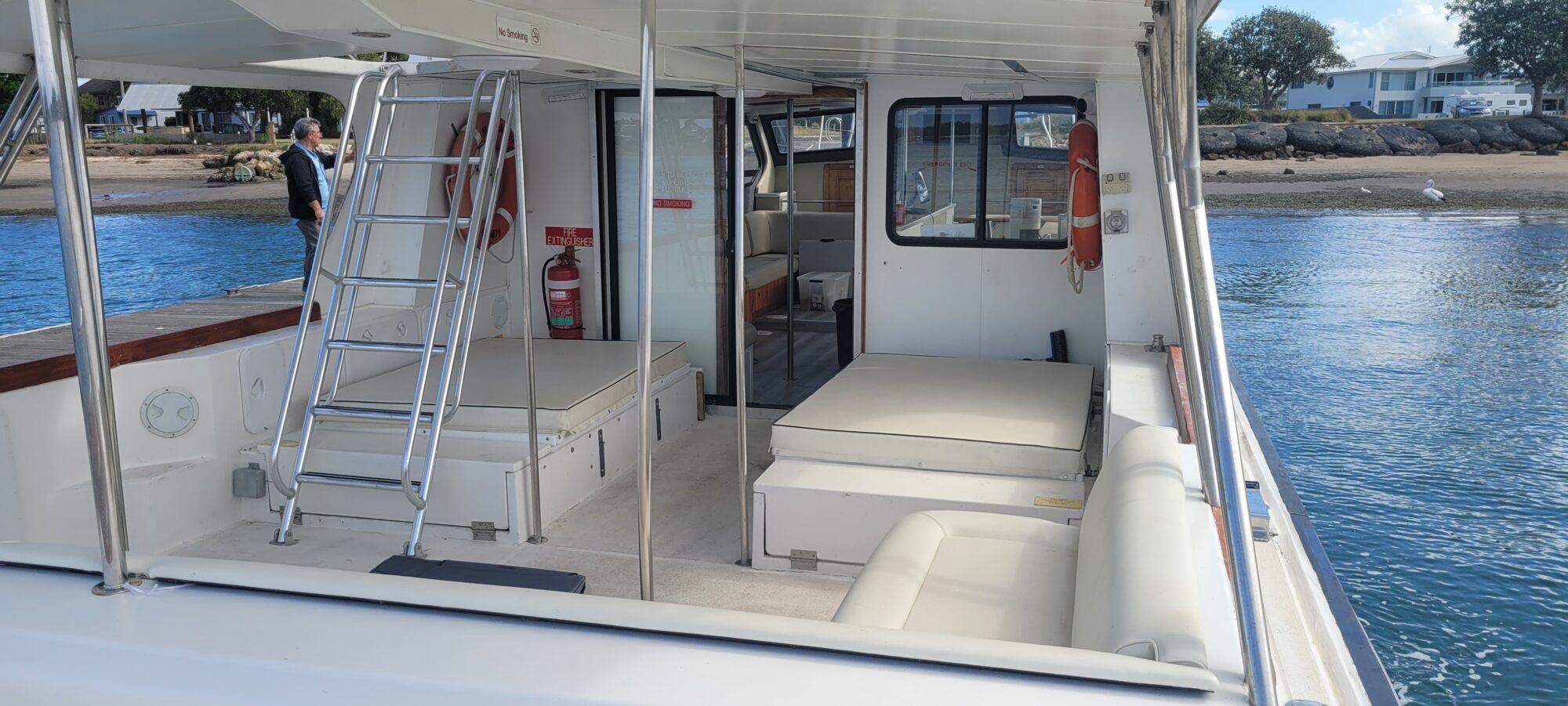 The photo shows the back deck of a white motorboat with cushioned seats, a metal ladder, and a fire extinguisher. The boat is docked near a shoreline with trees, rocks, and buildings visible in the background.