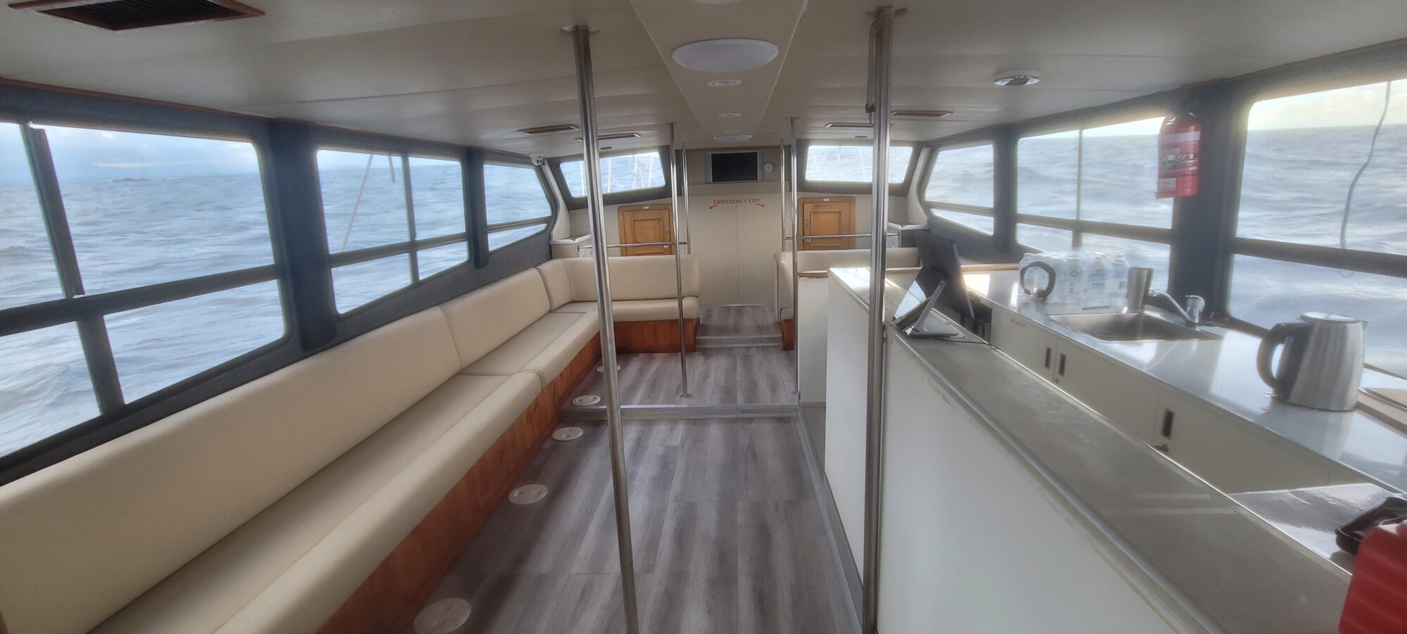 Interior of a boat with beige cushioned seating along the left side, windows showing choppy ocean waves, and a kitchenette area on the right with a kettle, mugs, and a sink.