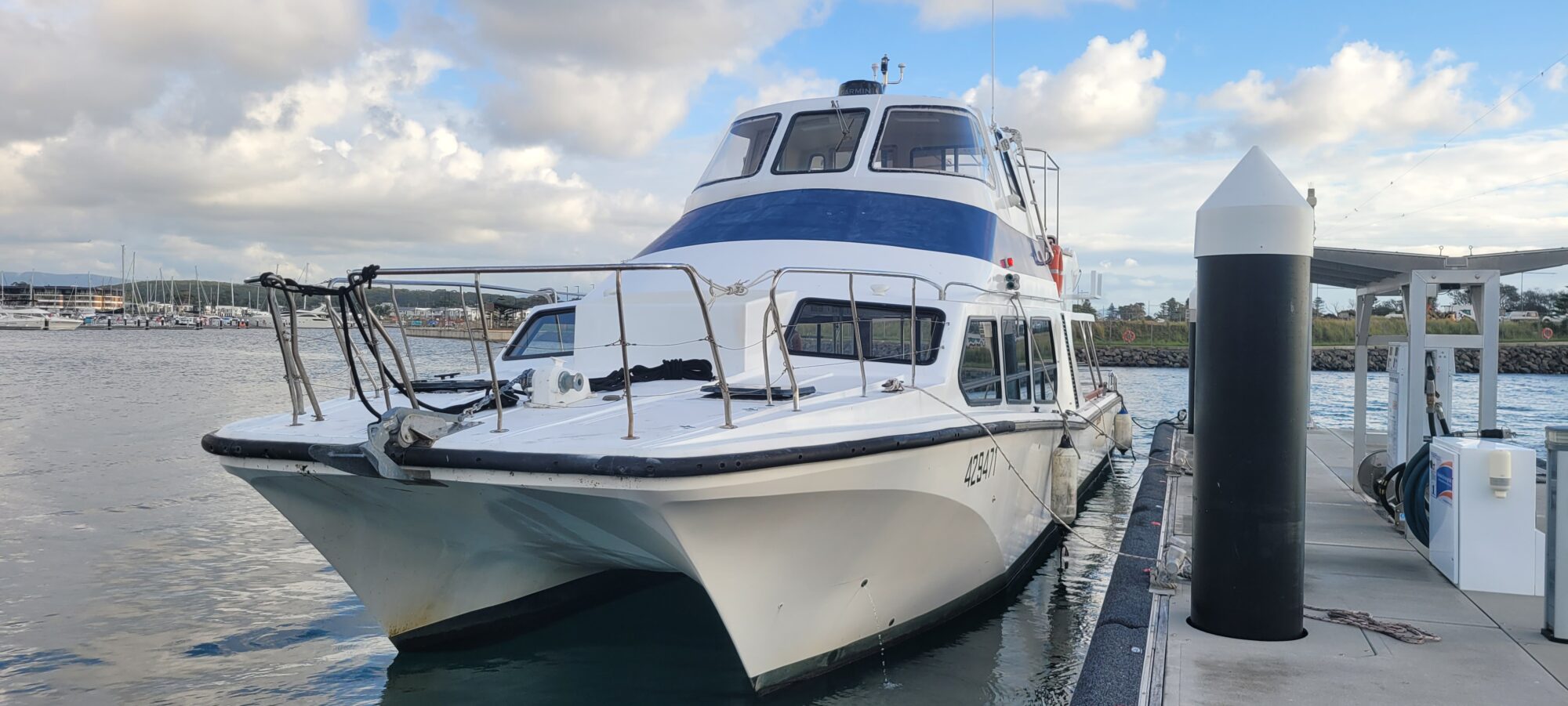 A white catamaran with two decks is docked at a marina. The boat is tied to a pier, with water and a cloudy sky in the background. Other boats and a shore are visible in the distance.