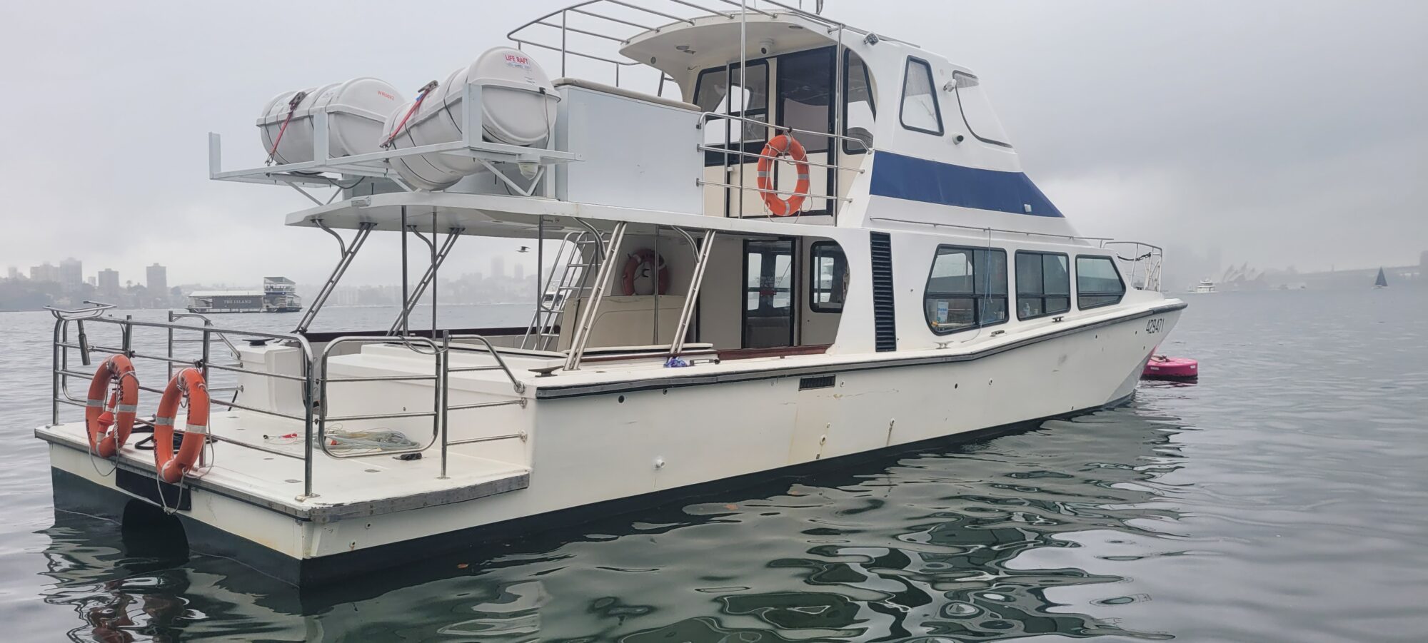 A white motor yacht with blue accents floats on calm water. It has orange life rings, life rafts on the upper deck, and a covered cabin. A cloudy sky and a distant city skyline are visible in the background.