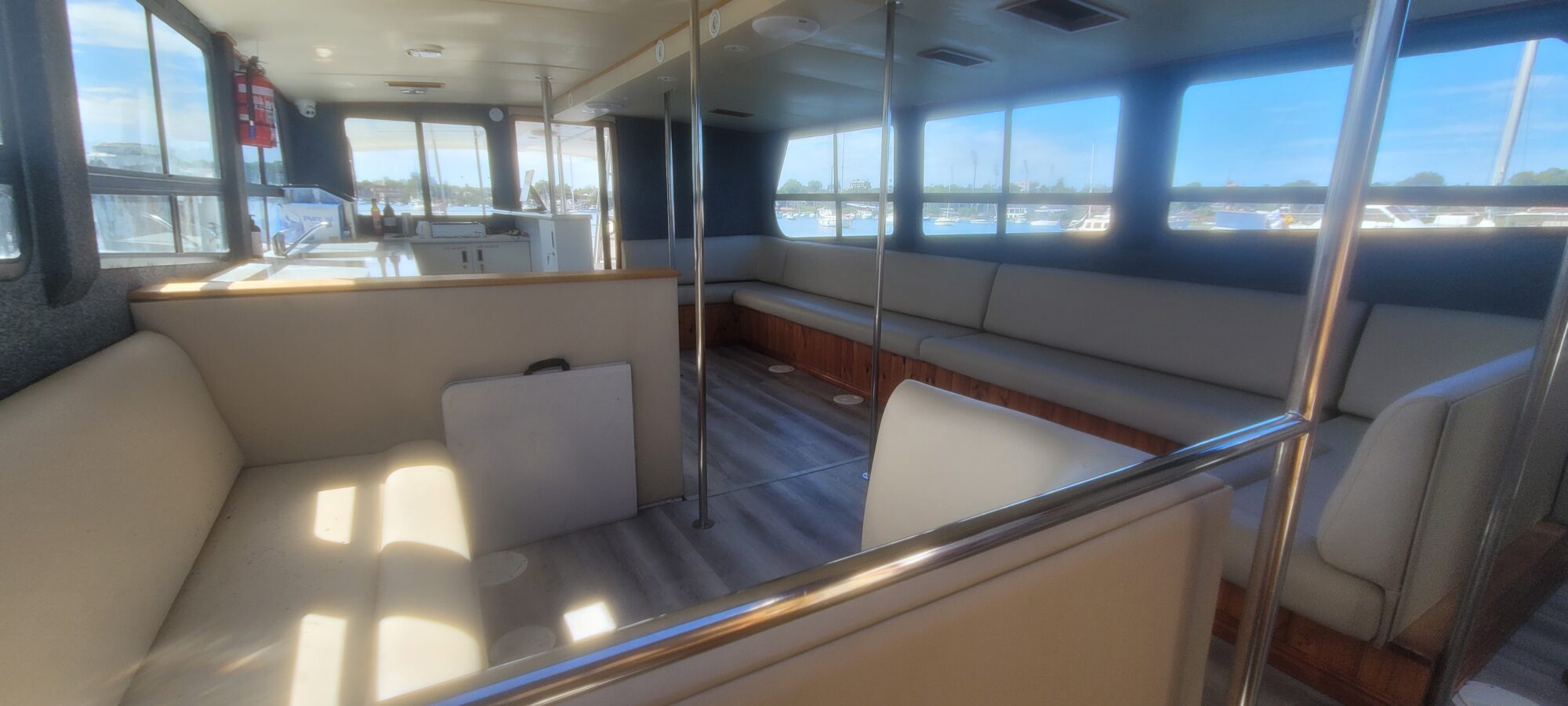 The image shows the interior of a boat with large windows, gray cushioned bench seating along the walls, metal poles, and light wood flooring. Sunlight streams in, and water and docks are visible outside.