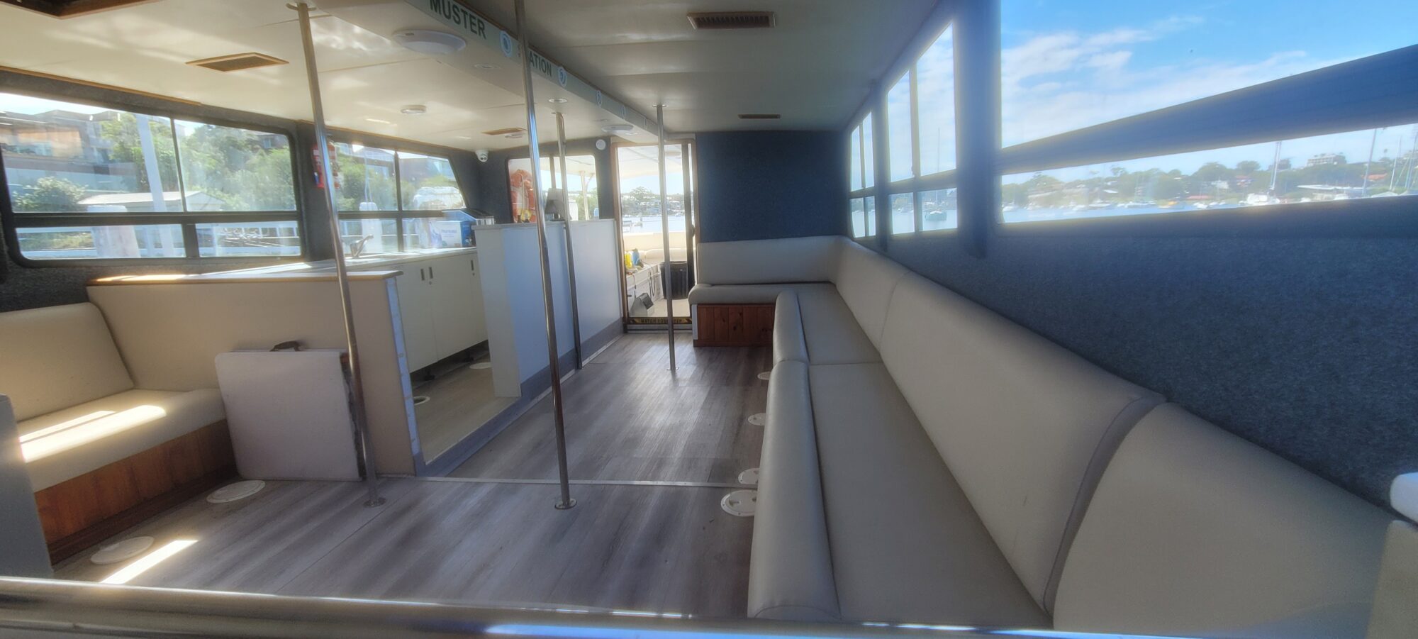 The image shows the interior of a boat with light-colored cushioned bench seating along the walls, large windows letting in sunlight, and a wood-look floor. The boat is docked, and the outside view shows cars and blue sky.