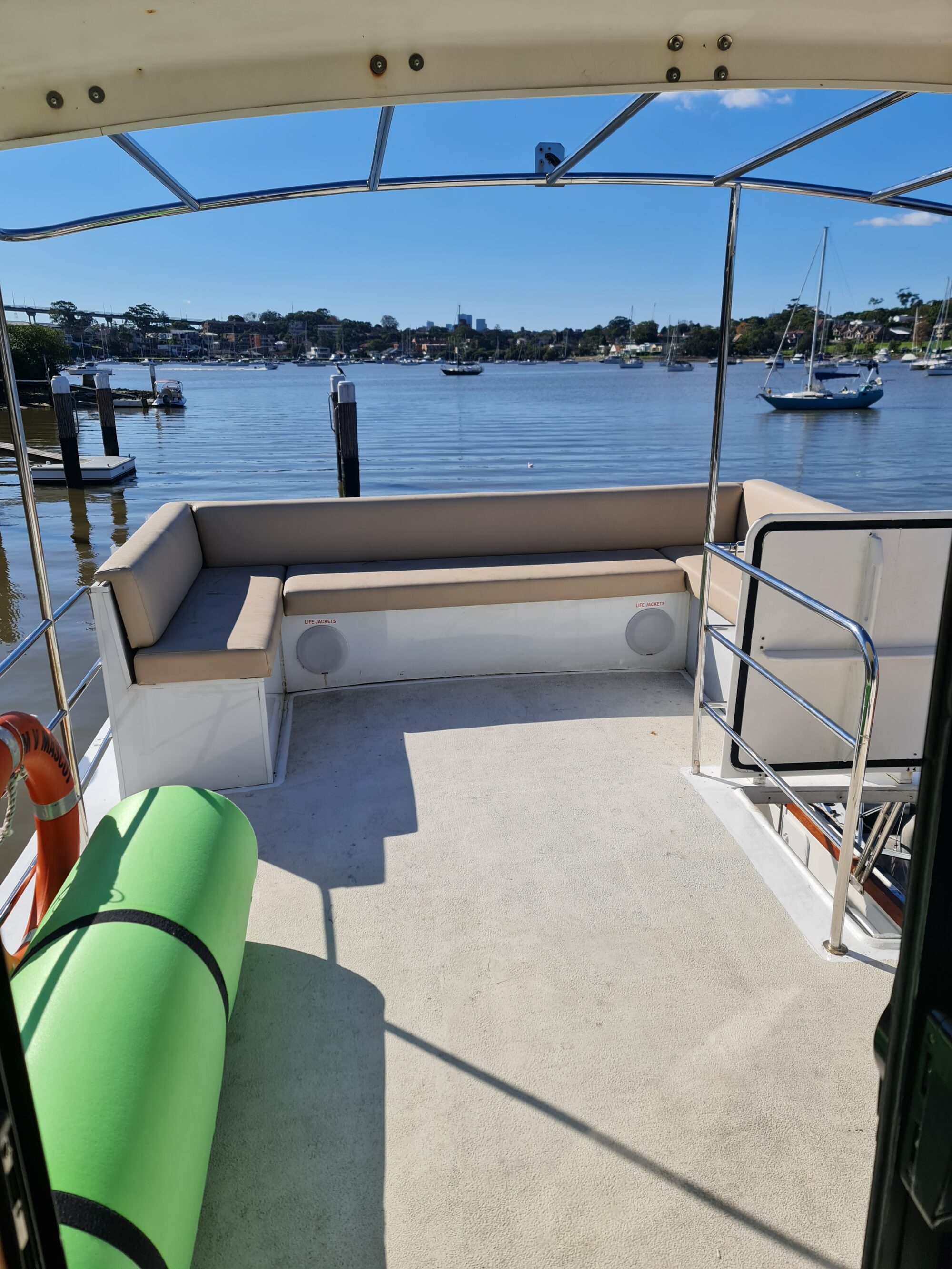 Sunny view from the covered back deck of a boat, showing a green yoga mat, tan bench seating, safety railing, and a calm harbor with sailboats and trees in the background.