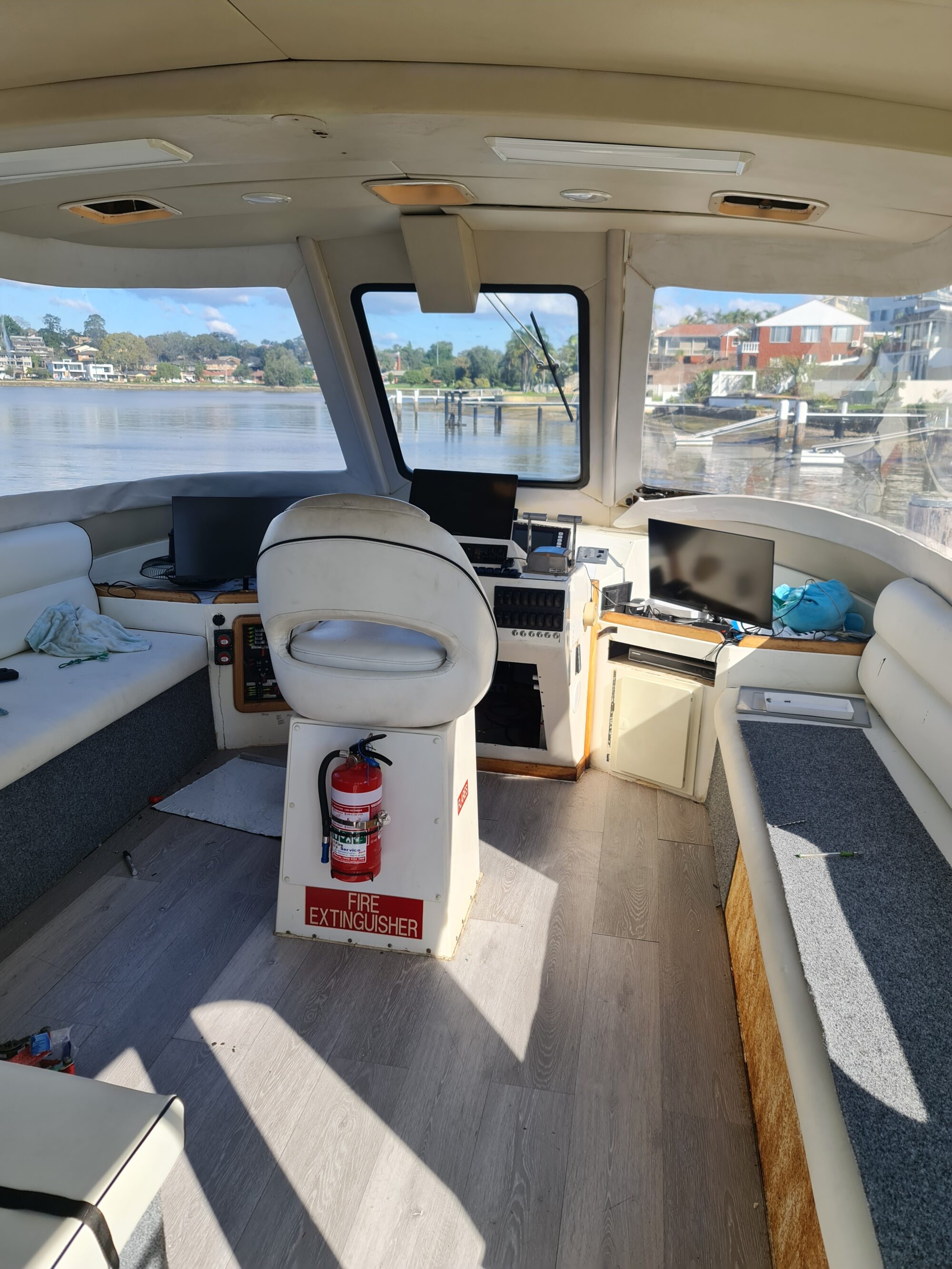 Interior of a boat’s control cabin with a central white captain’s chair, navigation screens, benches on both sides, large windows with a view of water and houses, and a fire extinguisher mounted at the chair’s base.
