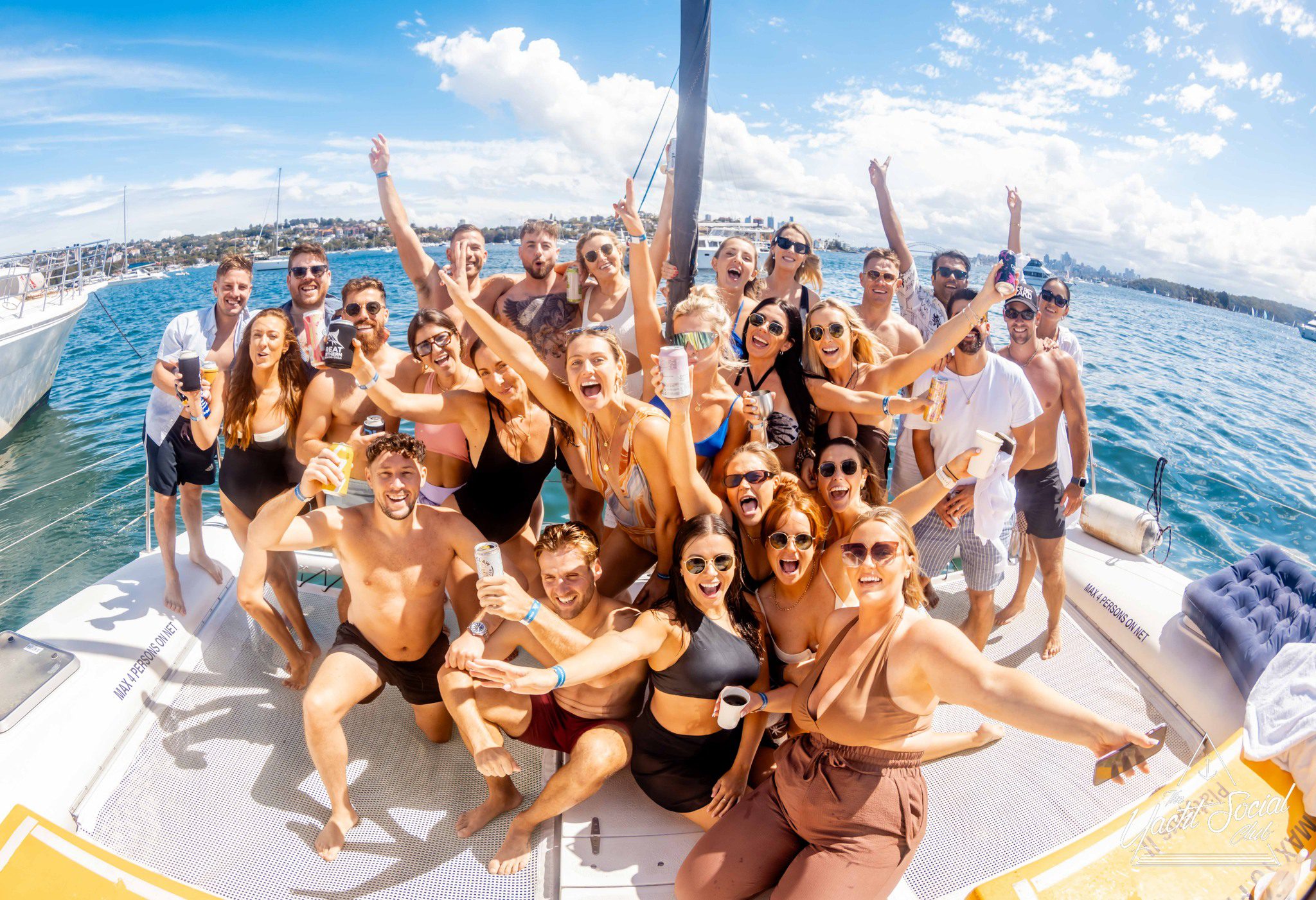 A large group of young adults in swimwear smile, cheer, and raise drinks while posing together on a boat during an Off The Apps yacht singles event, with blue water, other boats, and a city skyline in the background under a sunny sky.