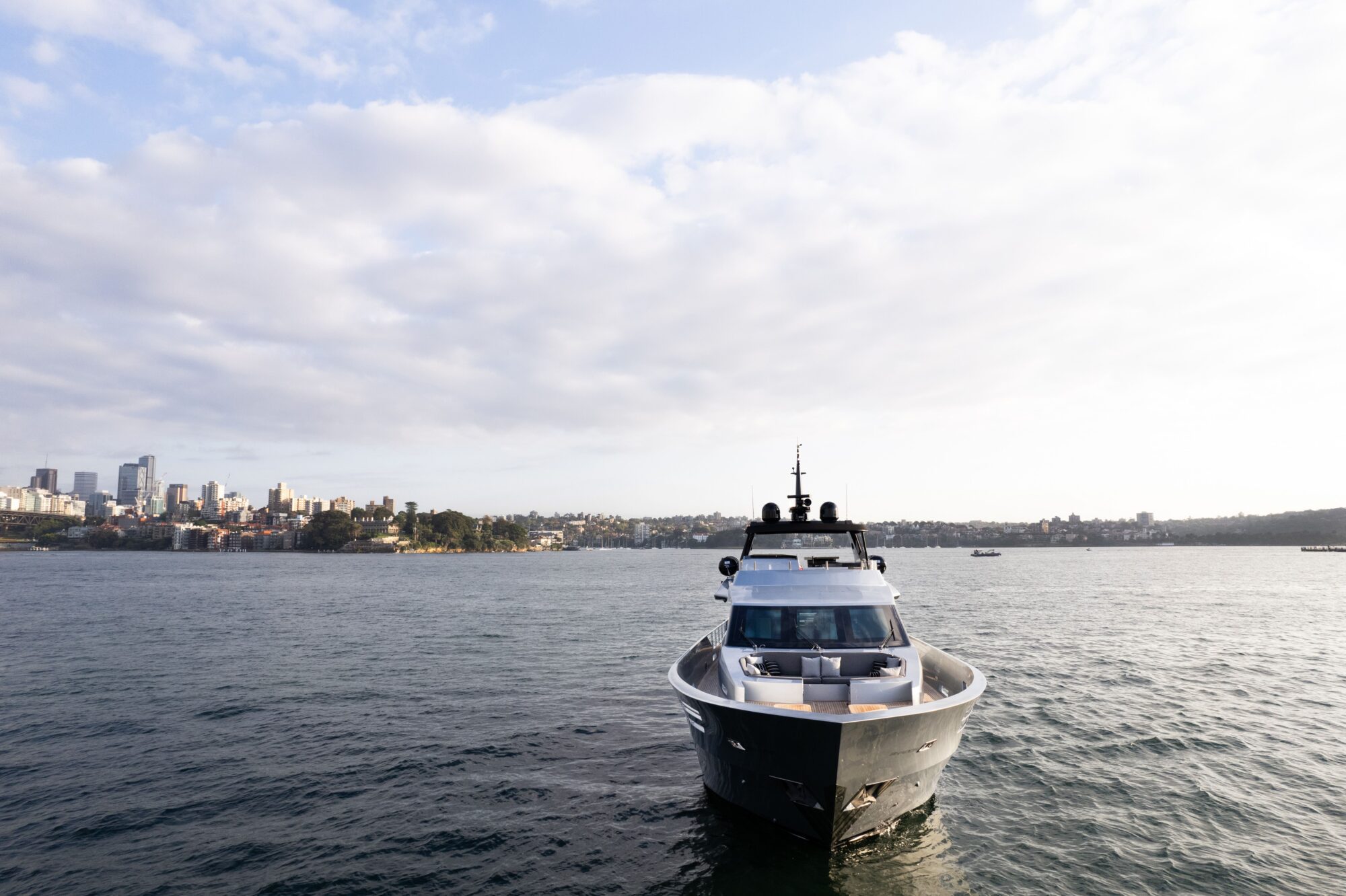 A modern yacht is anchored on a calm body of water with a city skyline and greenery visible in the distance under a partly cloudy sky.