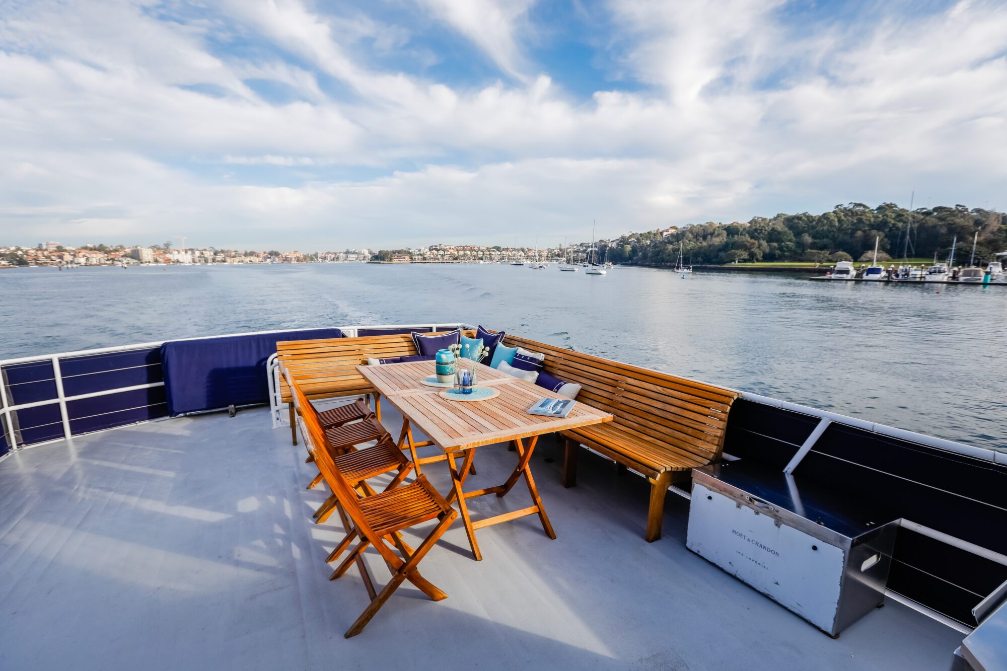 A wooden table with chairs and benches is set on the deck of a boat, overlooking calm water with sailboats and a tree-lined shoreline in the background under a partly cloudy sky.