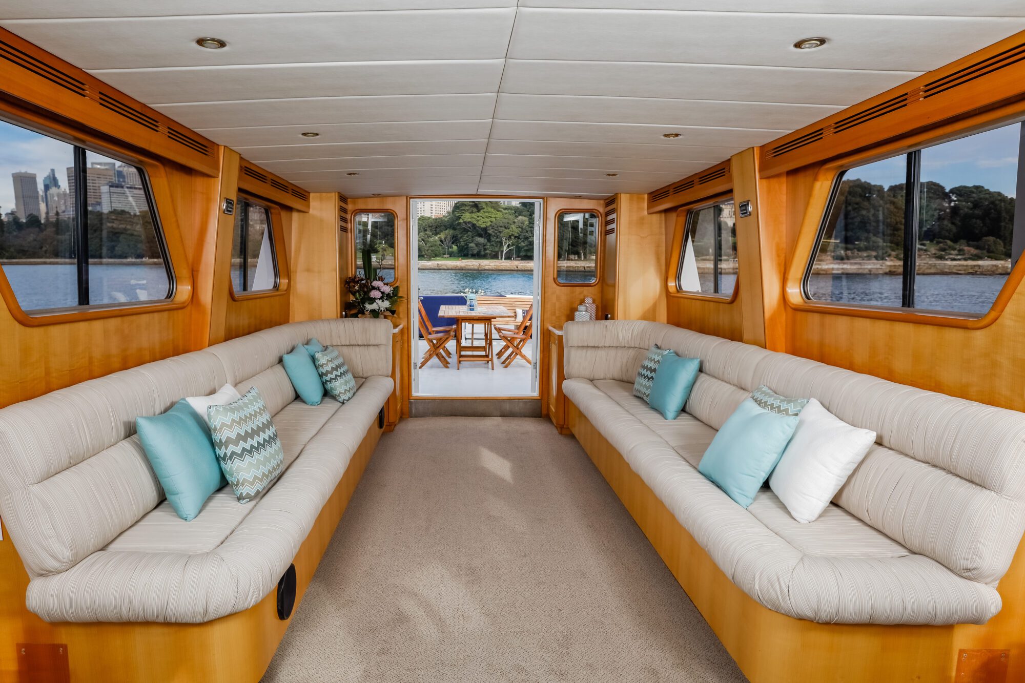 The interior of a boat with two long beige sofas lined with blue and white pillows, large windows on both sides, and a view of a table and chairs on an outdoor deck overlooking water and greenery.