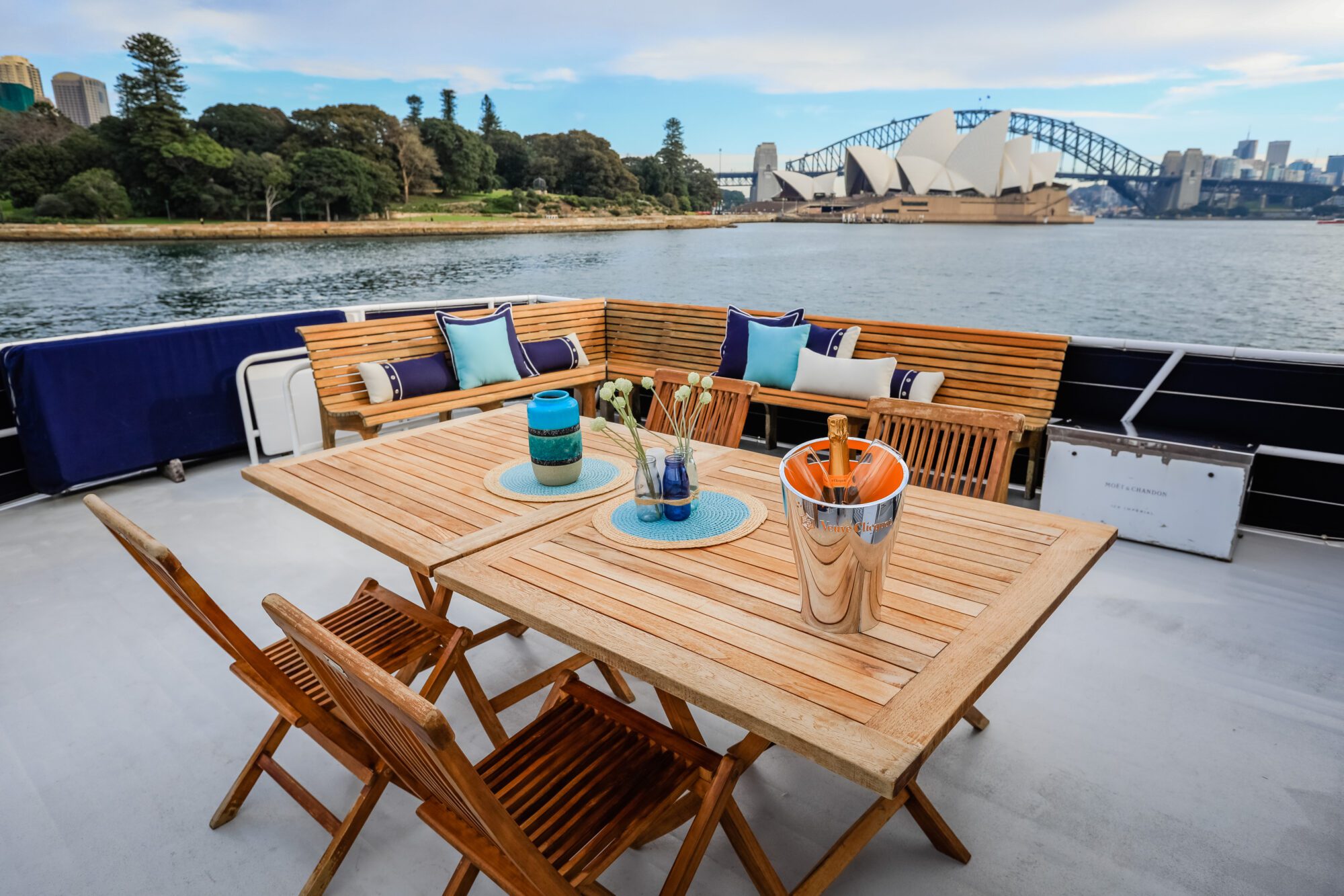 A wooden outdoor dining set and bench with colorful cushions on a boat deck overlooks Sydney Opera House and Harbour Bridge across the water on a clear day.