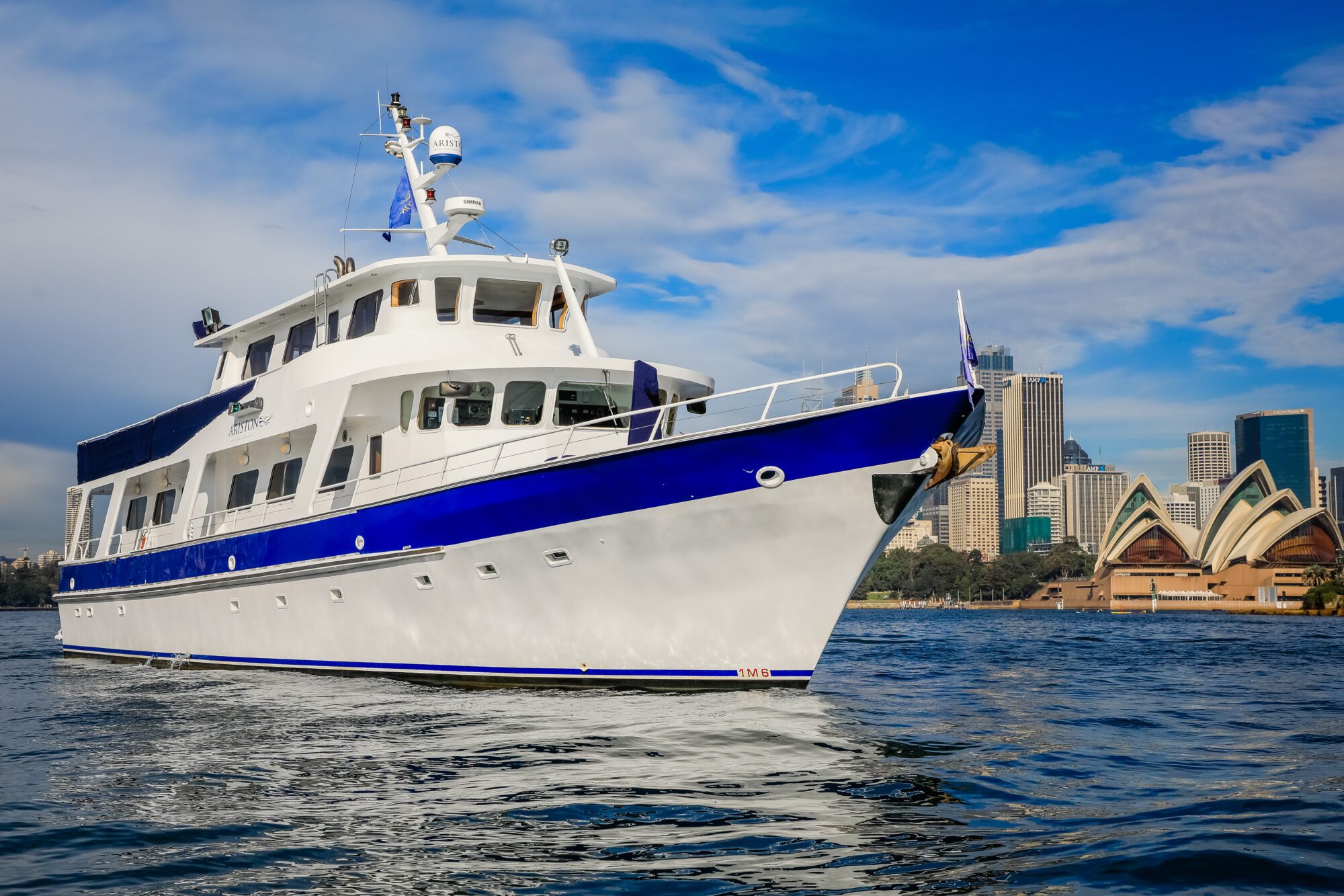 A white and blue yacht is sailing on calm water with a city skyline and the Sydney Opera House visible in the background under a partly cloudy sky.