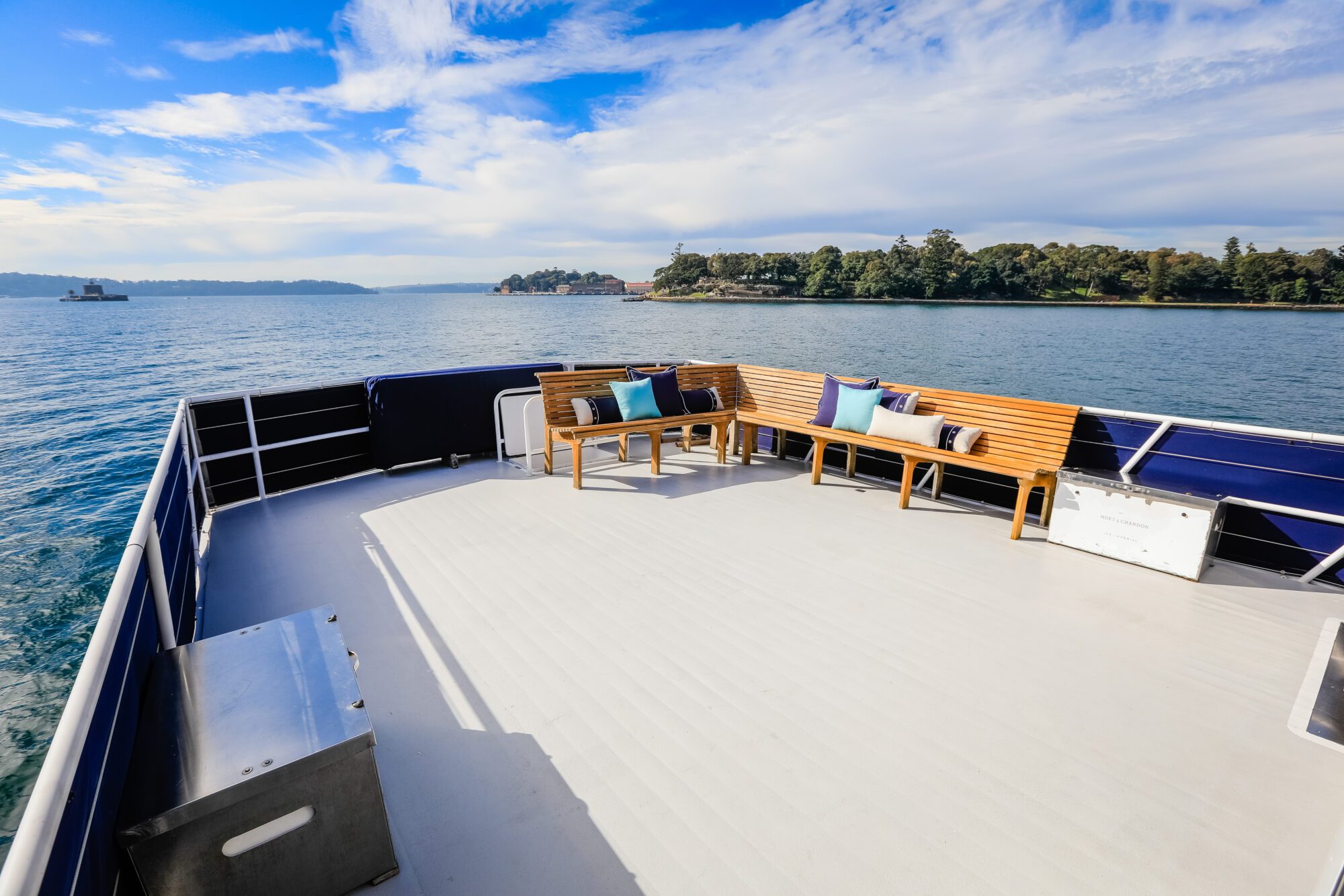 The image shows the open deck of a boat with wooden benches and blue cushions, overlooking calm water and green shoreline under a bright, partly cloudy sky.