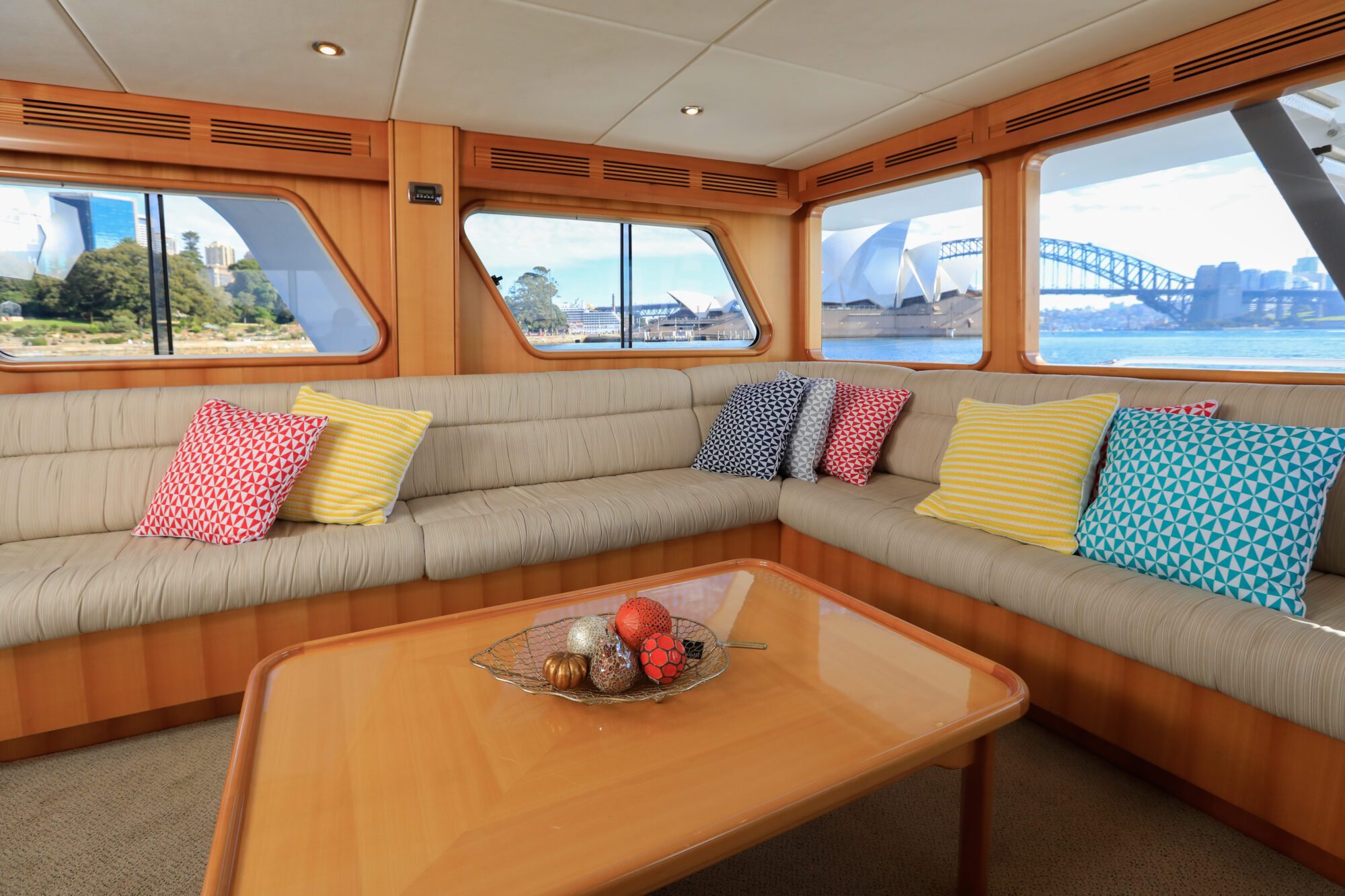 The interior of a boat with beige cushioned seating, colorful patterned pillows, and a wooden table with decorative balls. Large windows offer views of the Sydney Opera House and Harbour Bridge.