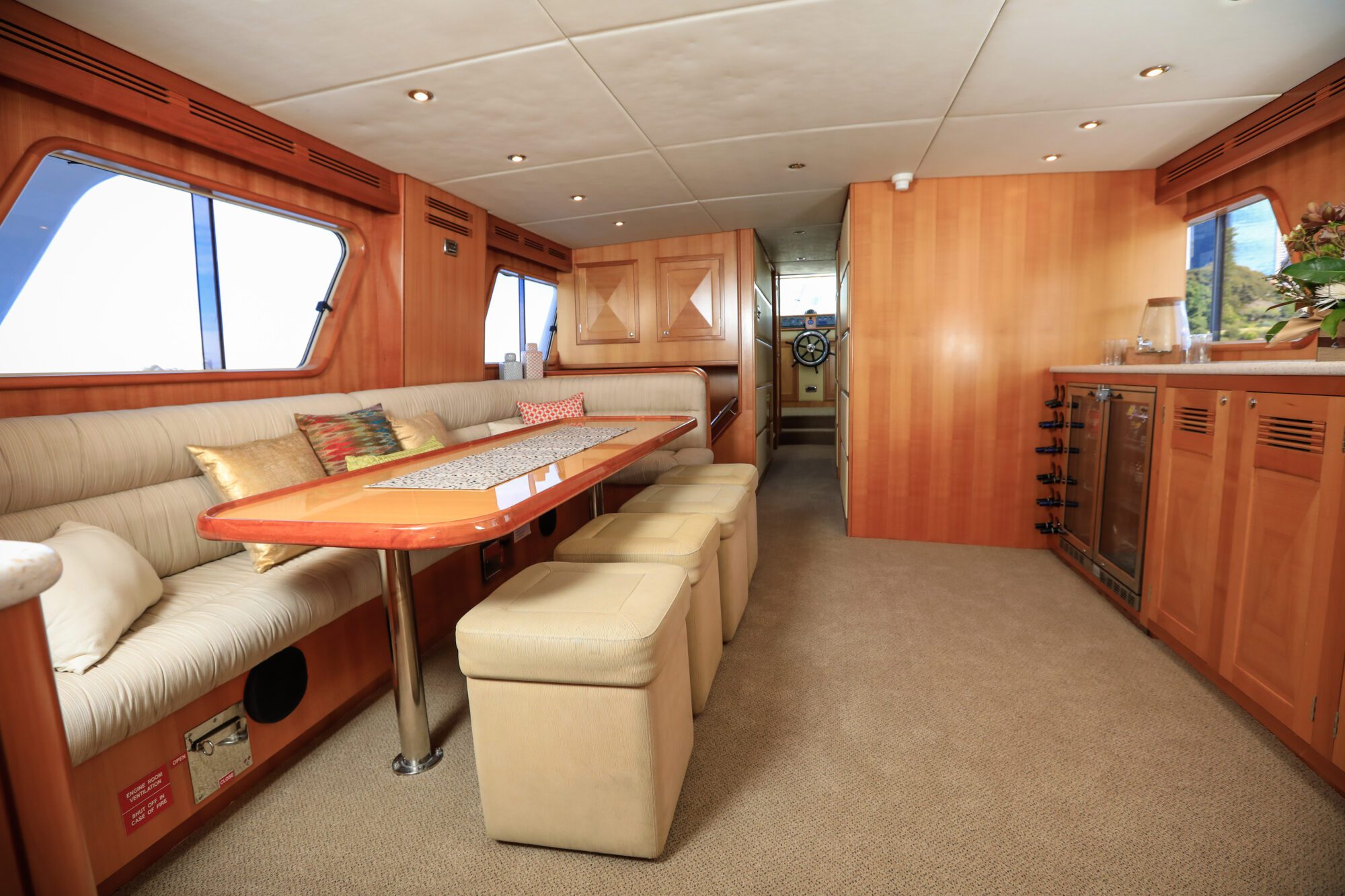 Interior of a yacht with a beige sofa and cushions, a long dining table with padded stools, wood paneling, carpeted floor, and a small kitchen area; a steering wheel is visible at the far end.