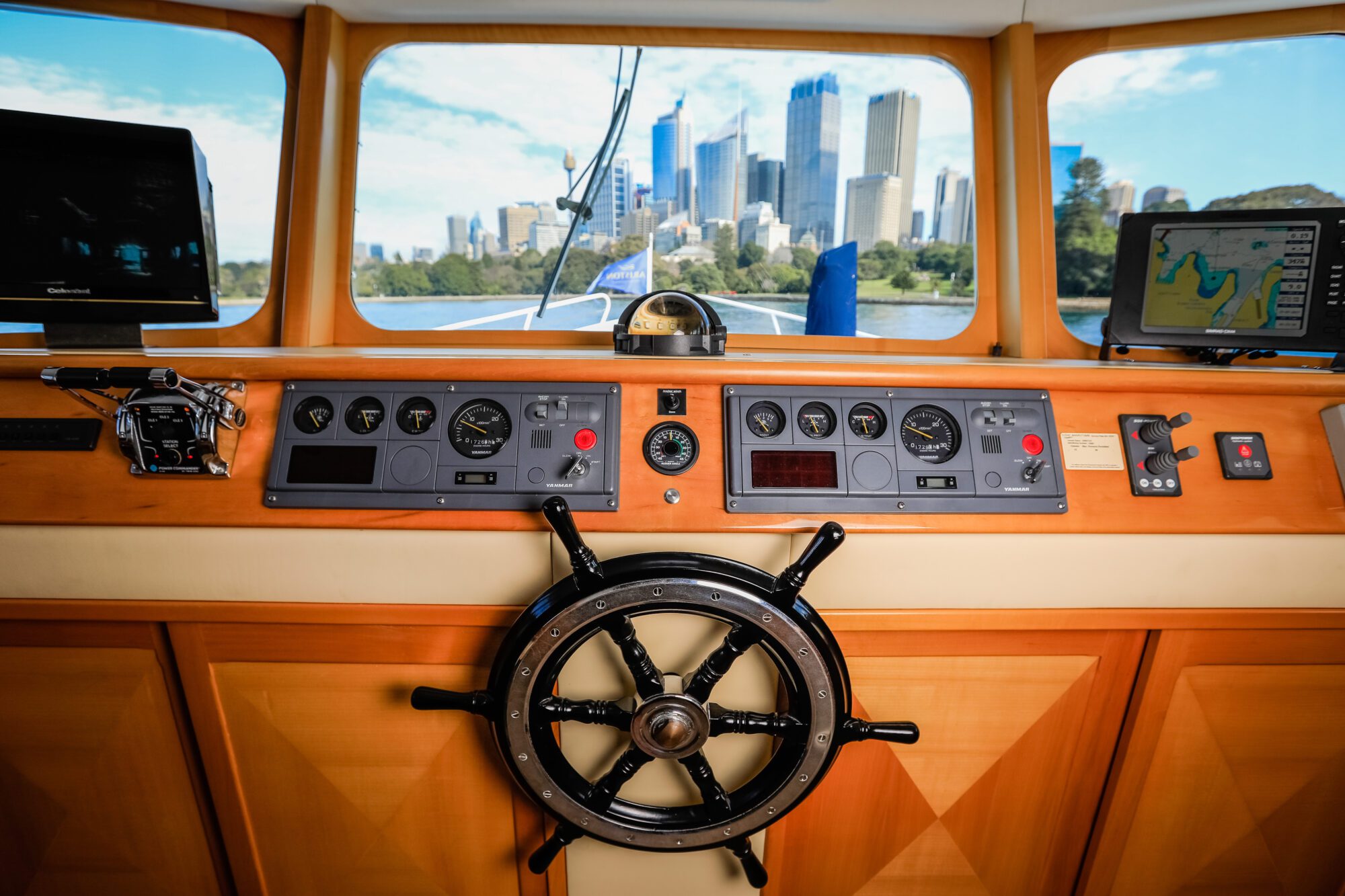 View from a boat's helm with a large steering wheel, control panels, and navigation screens. Through the windows, a city skyline with tall buildings and a clear blue sky is visible.