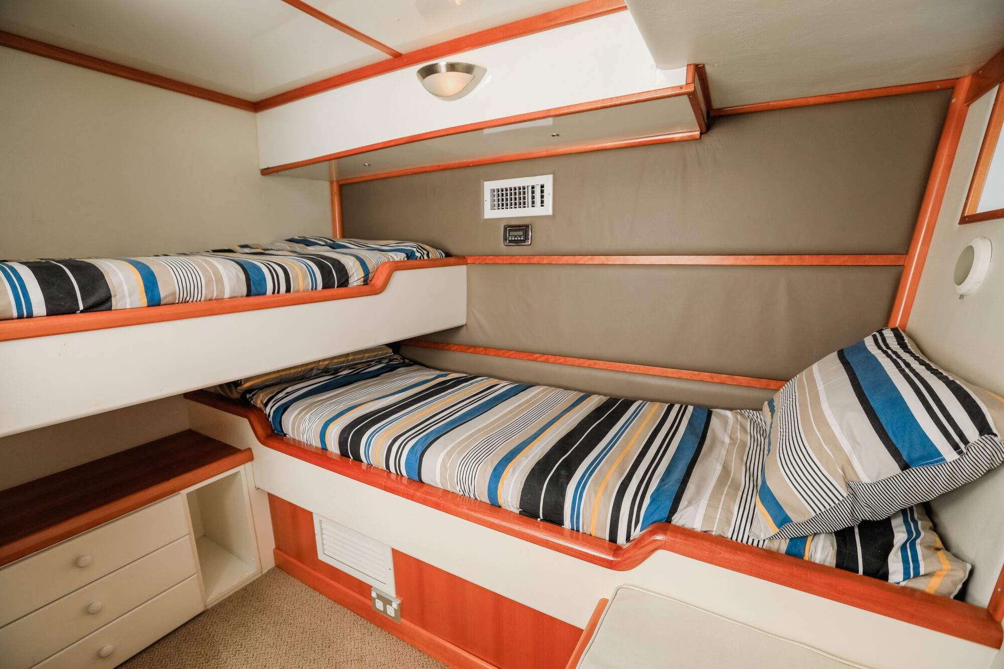 Small room with two striped bedding bunk beds, one above the other. There are light-colored walls, wooden trim, a small drawer unit, and a built-in shelf. The space appears neat and compact.