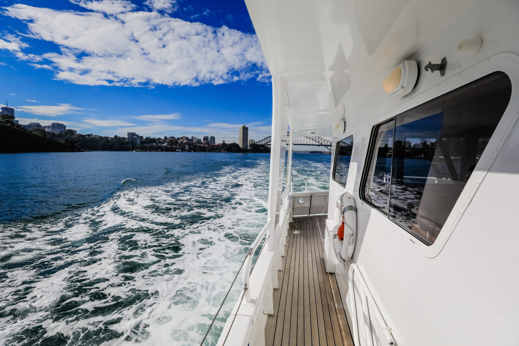 View from the side deck of a boat moving through blue water, leaving a foamy wake behind. City buildings, a bridge, and green shoreline are visible under a partly cloudy sky.