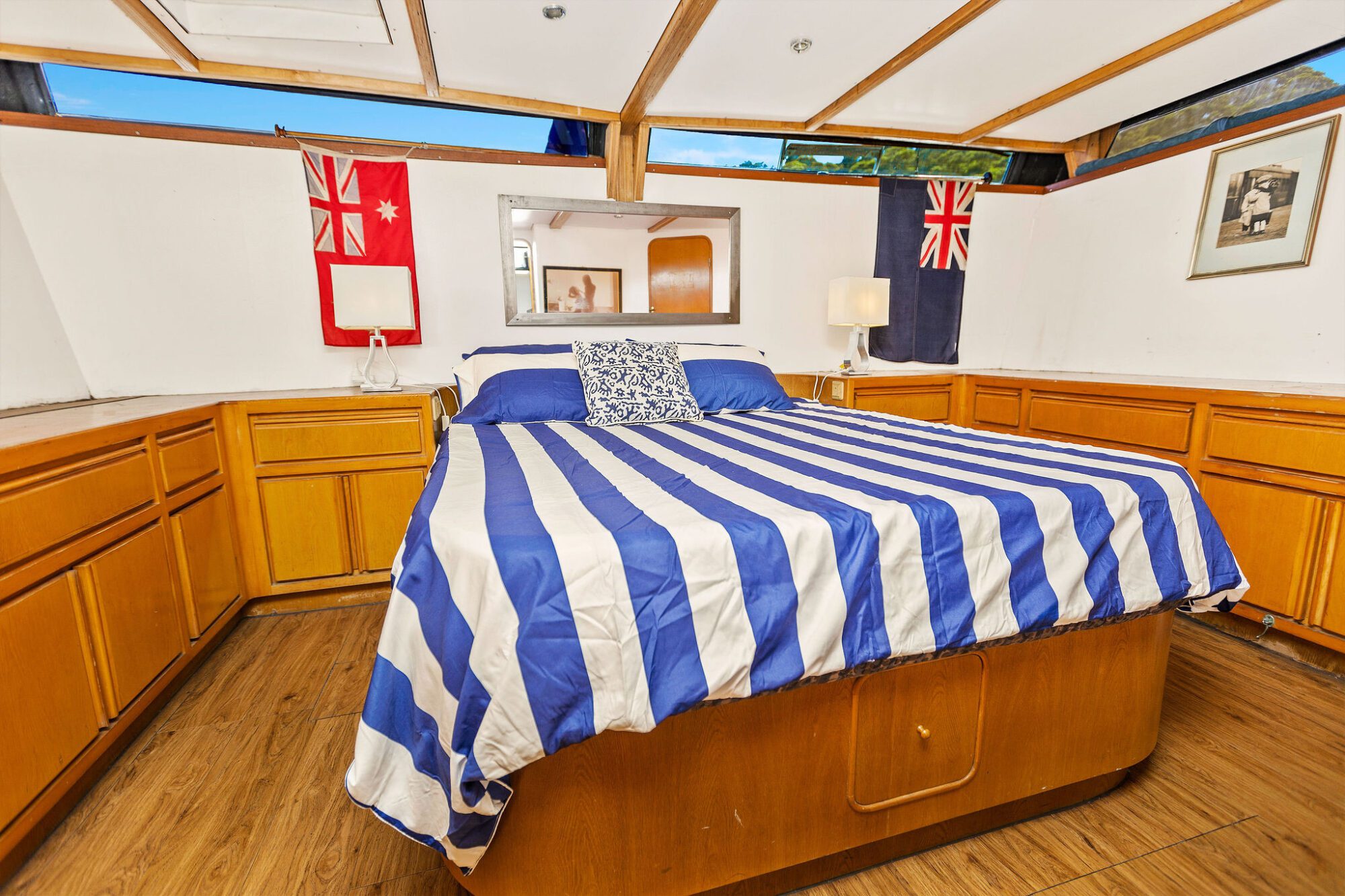 A bedroom with wood-paneled cabinets, a bed with a blue and white striped bedspread, two nautical flags on the walls, two table lamps, and a framed picture above the headboard.
