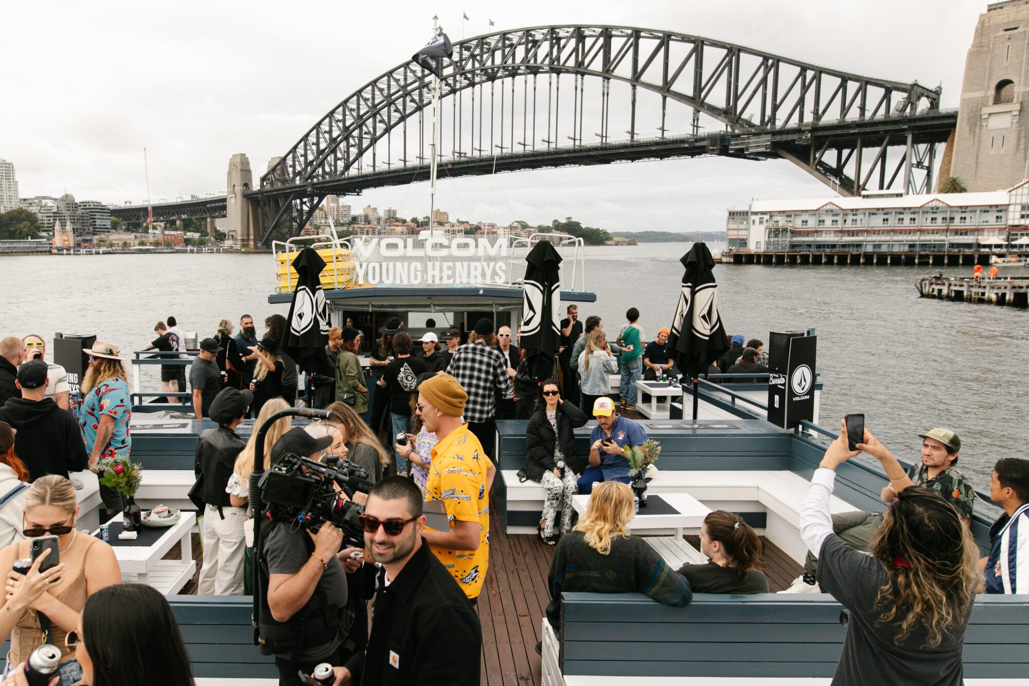 A group of people socializing on a boat with picnic tables, set against the backdrop of Sydney Harbour Bridge on a cloudy day. Signs on the boat read “Volcom Young Henrys.” Some guests are taking photos and filming.