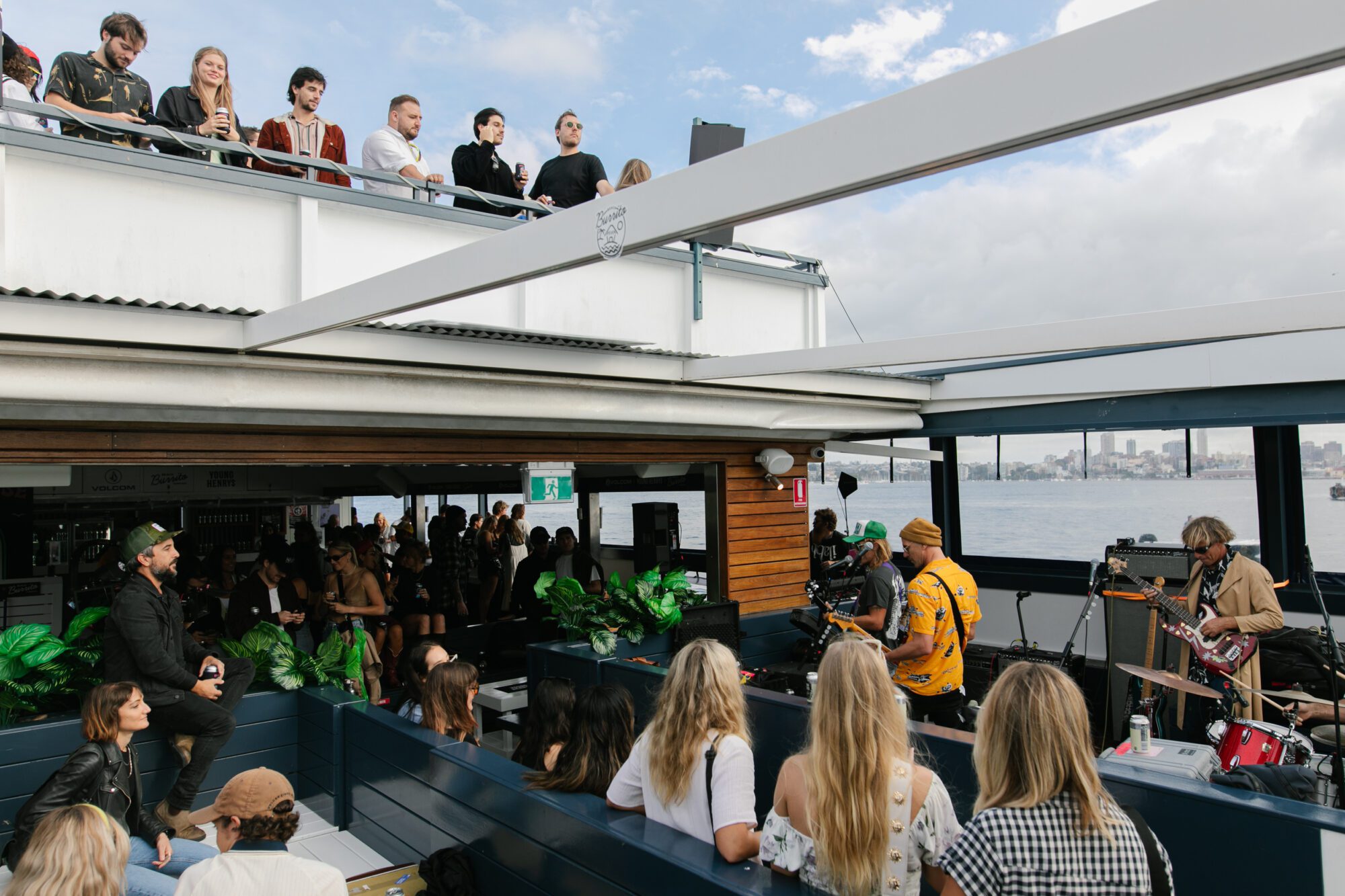 A band performs live on an outdoor rooftop venue by the water, with people watching from both the deck and an upper balcony under a partly cloudy sky.