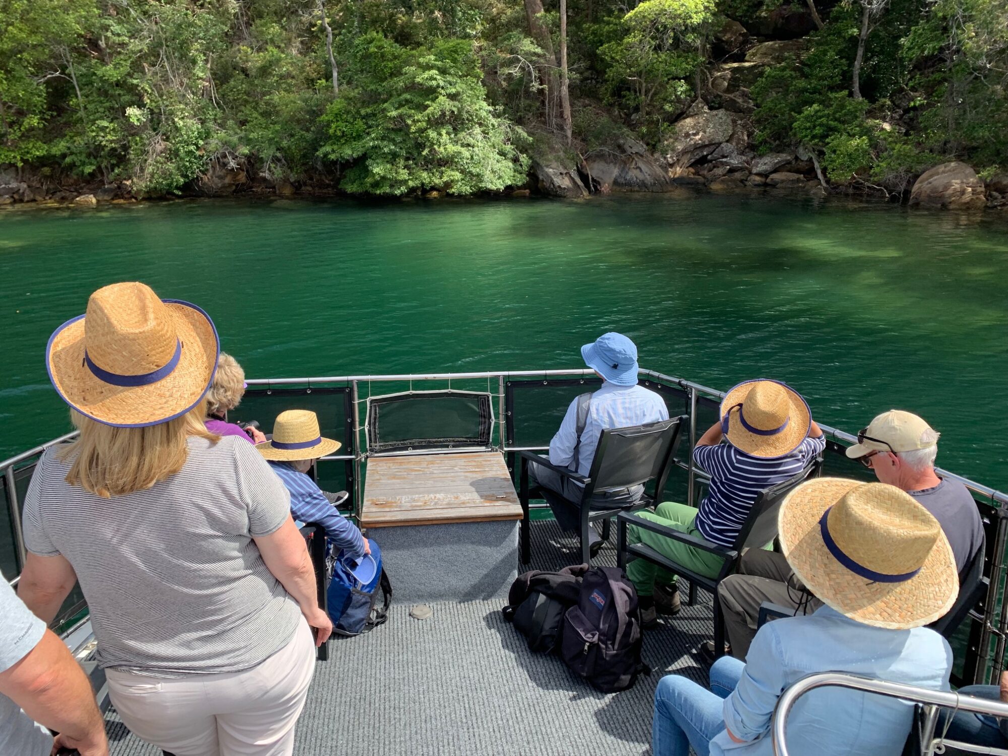 A group of people wearing straw hats sit and stand on a boat deck, facing green water and lush forested shoreline. Some have backpacks at their feet and the weather appears sunny.