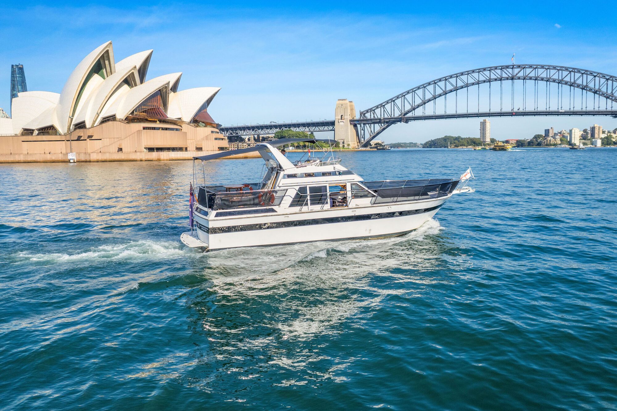 A white motorboat cruises on blue water with the Sydney Opera House and Sydney Harbour Bridge in the background on a sunny day.