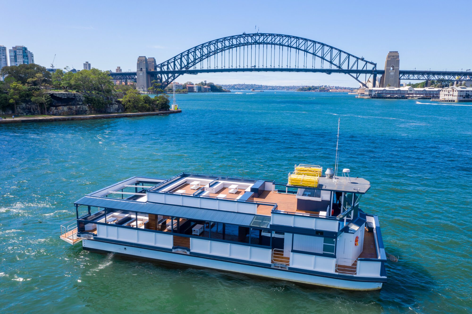 A modern houseboat floats on blue water with the Sydney Harbour Bridge in the background under a clear sky, and city buildings and greenery visible along the shoreline.