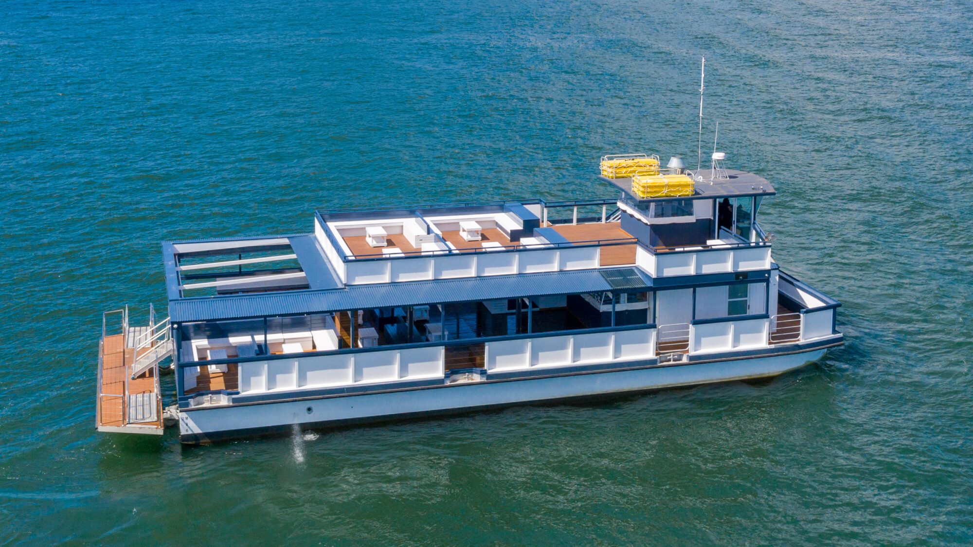 A modern white and blue two-level boat with open seating areas and lifeboats on the roof floats on calm blue water. The boat has large windows and an outdoor deck at the stern.