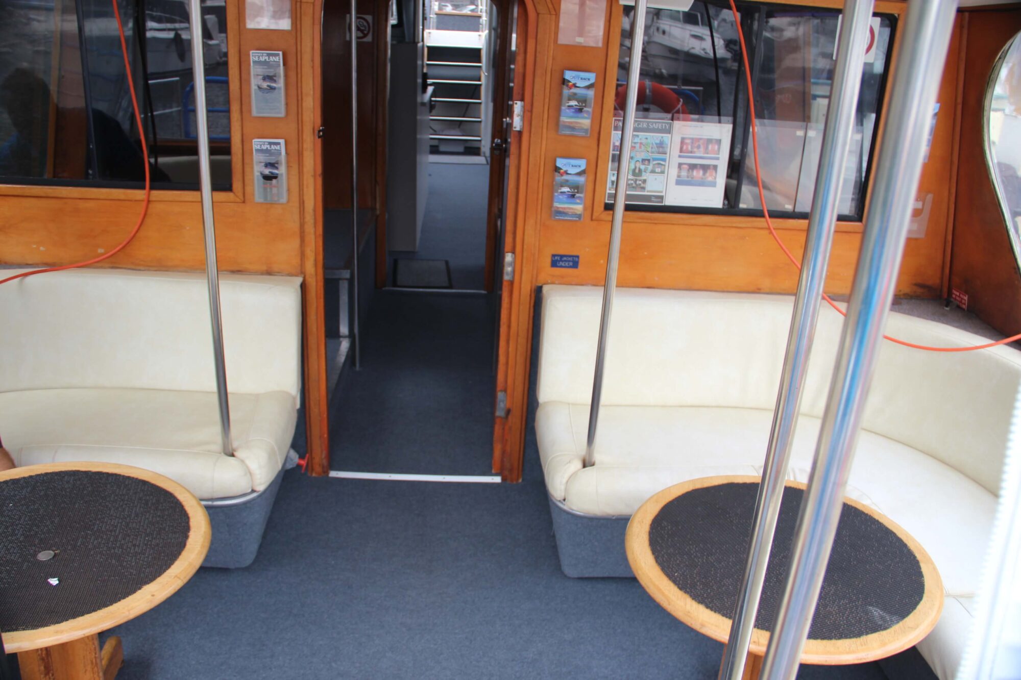 Interior of a boat with two round tables, cream-colored curved bench seating, metal poles, carpeted floor, wooden walls, and a doorway leading to stairs at the back. Posters and windows are visible on the walls.