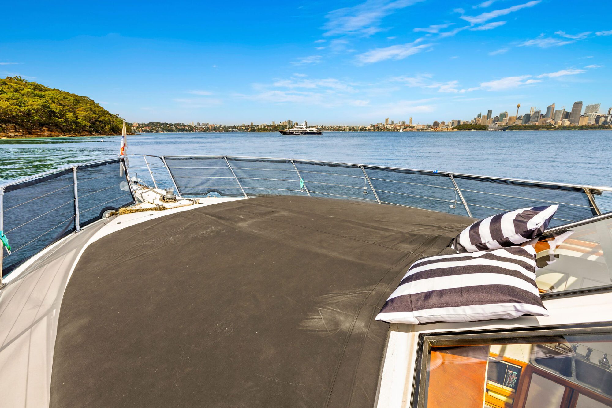 View from the bow of a boat with a striped pillow, overlooking calm water, city skyline, a distant ferry, and greenery under a clear blue sky.