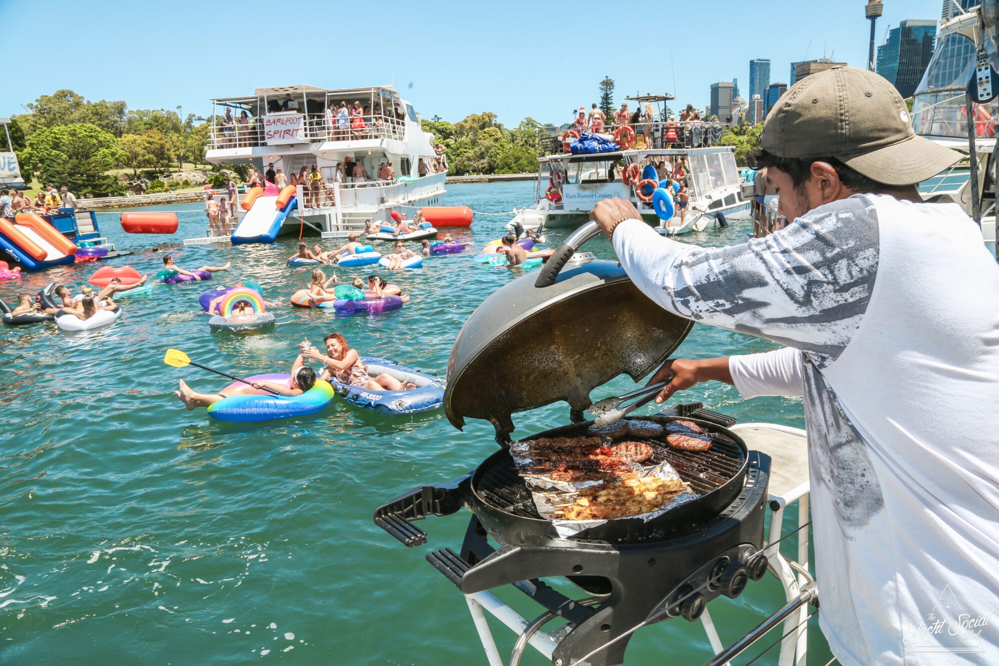 A man grills burgers on a boat, surrounded by people in colorful floats and inflatables enjoying a lively party on the water. Other boats with more partygoers are anchored nearby under a clear, sunny sky.