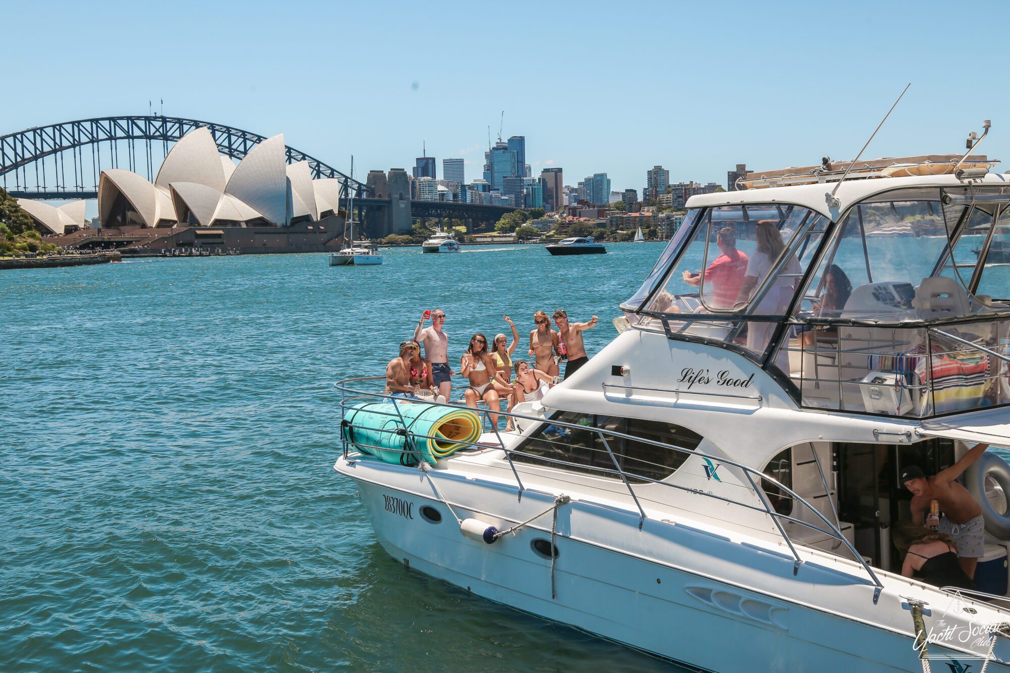 A group of people on a white yacht wave and pose for a photo in Sydney Harbour, with the Sydney Opera House and Harbour Bridge visible in the background on a sunny day.