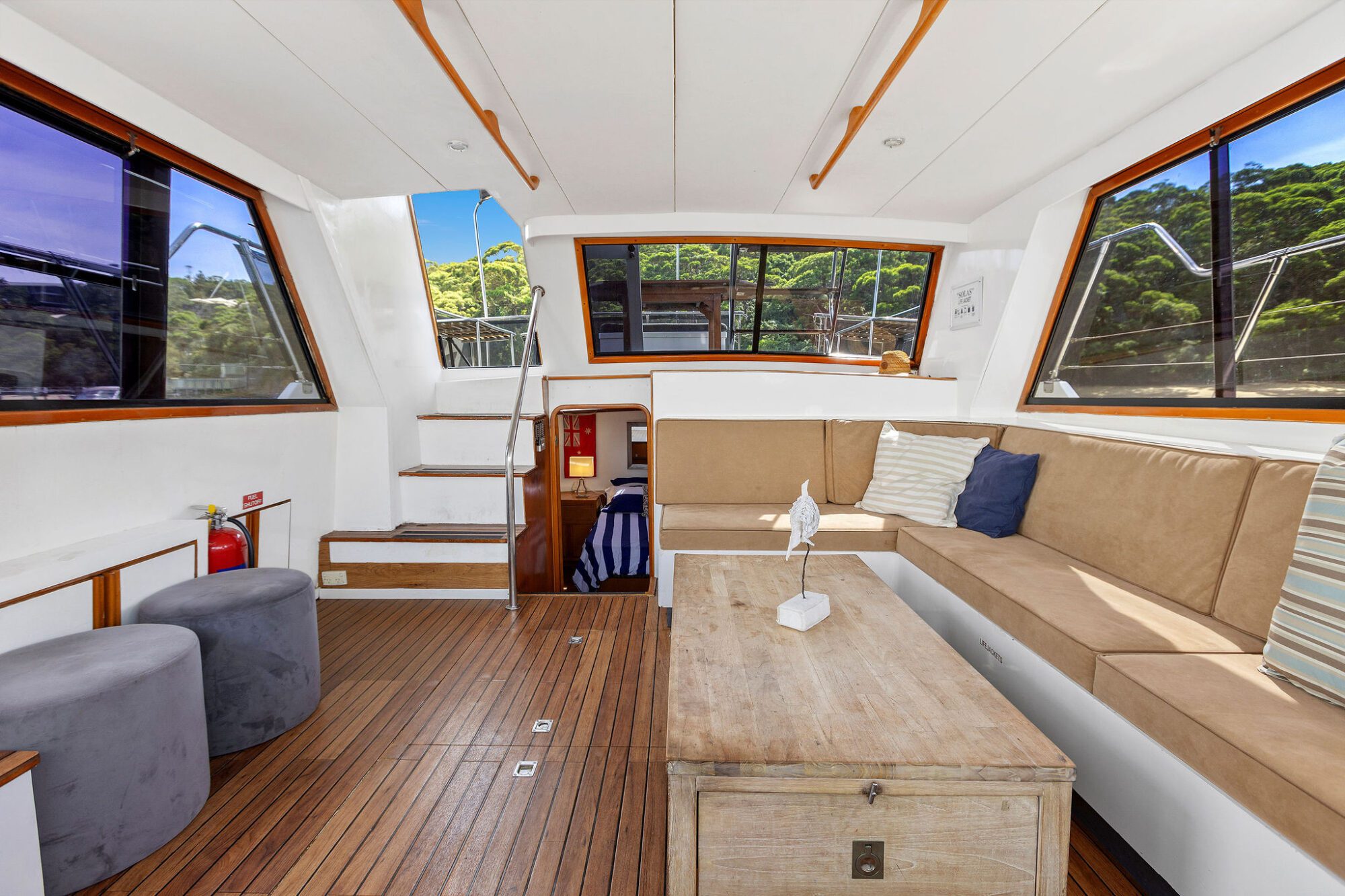 Interior of a boat cabin featuring tan cushioned seating, a wooden table, large windows with views of trees, stairs leading to a lower level, and light wood flooring. Pillows and decor create a cozy, inviting atmosphere.