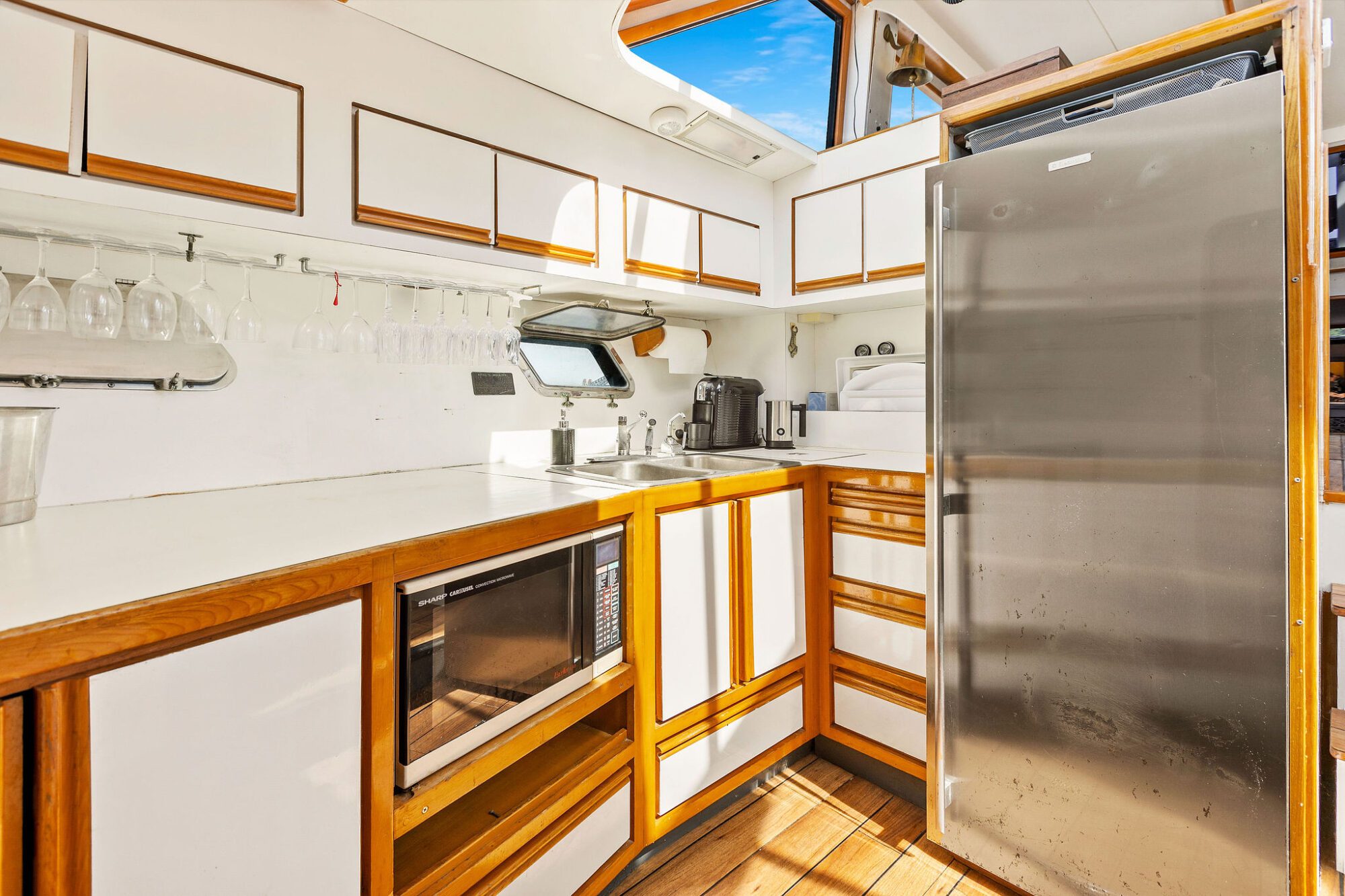 A compact, modern boat kitchen with white cabinets, wood trim, a stainless steel refrigerator, a microwave, a coffee maker, hanging wine glasses, and a small sink beneath a window. Sunlight streams in from a hatch above.