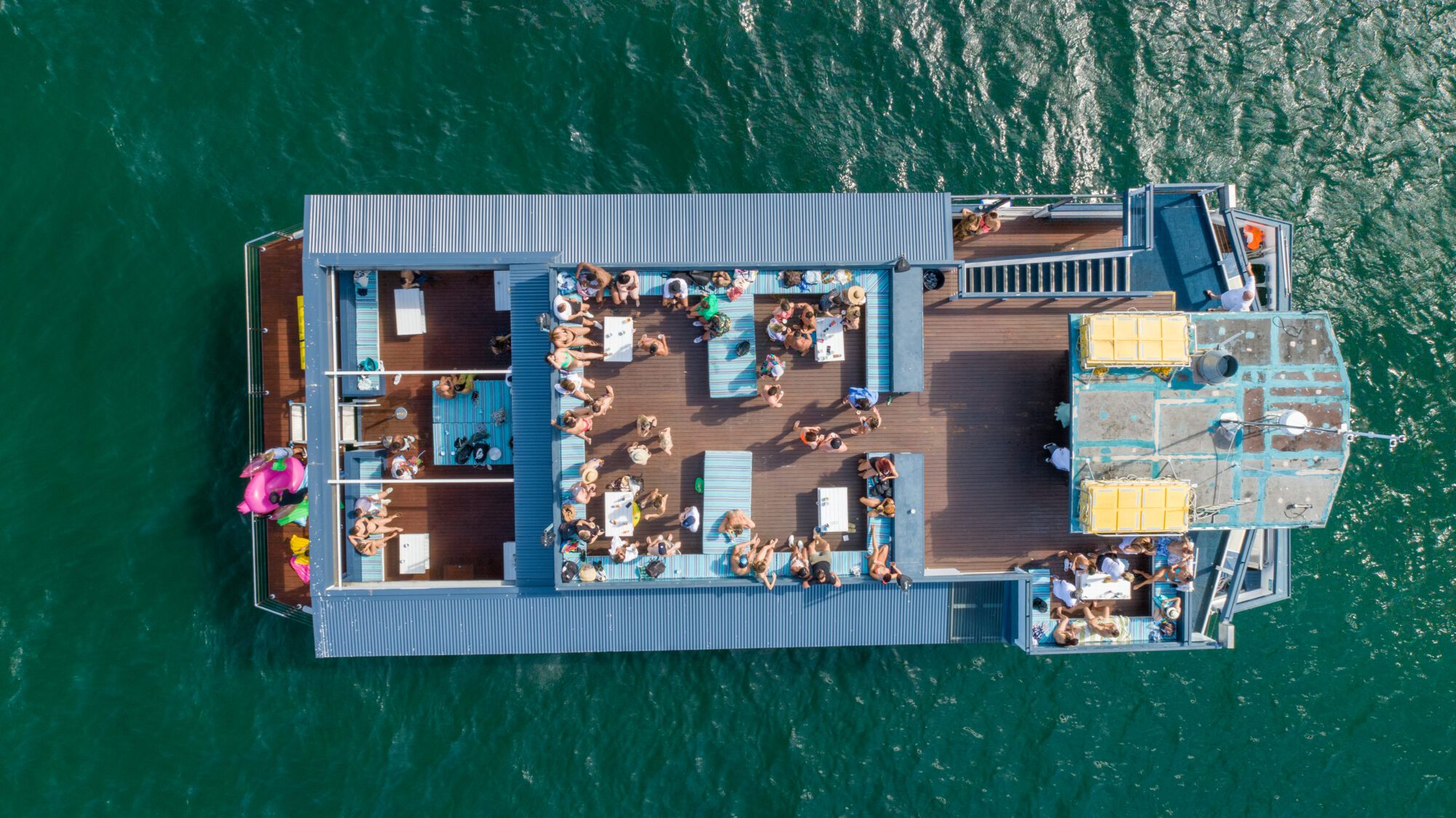 Aerial view of a large boat on water with many people on deck lounging, socializing, and enjoying the sunny weather. The boat has multiple seating areas and a visible pink inflatable float.