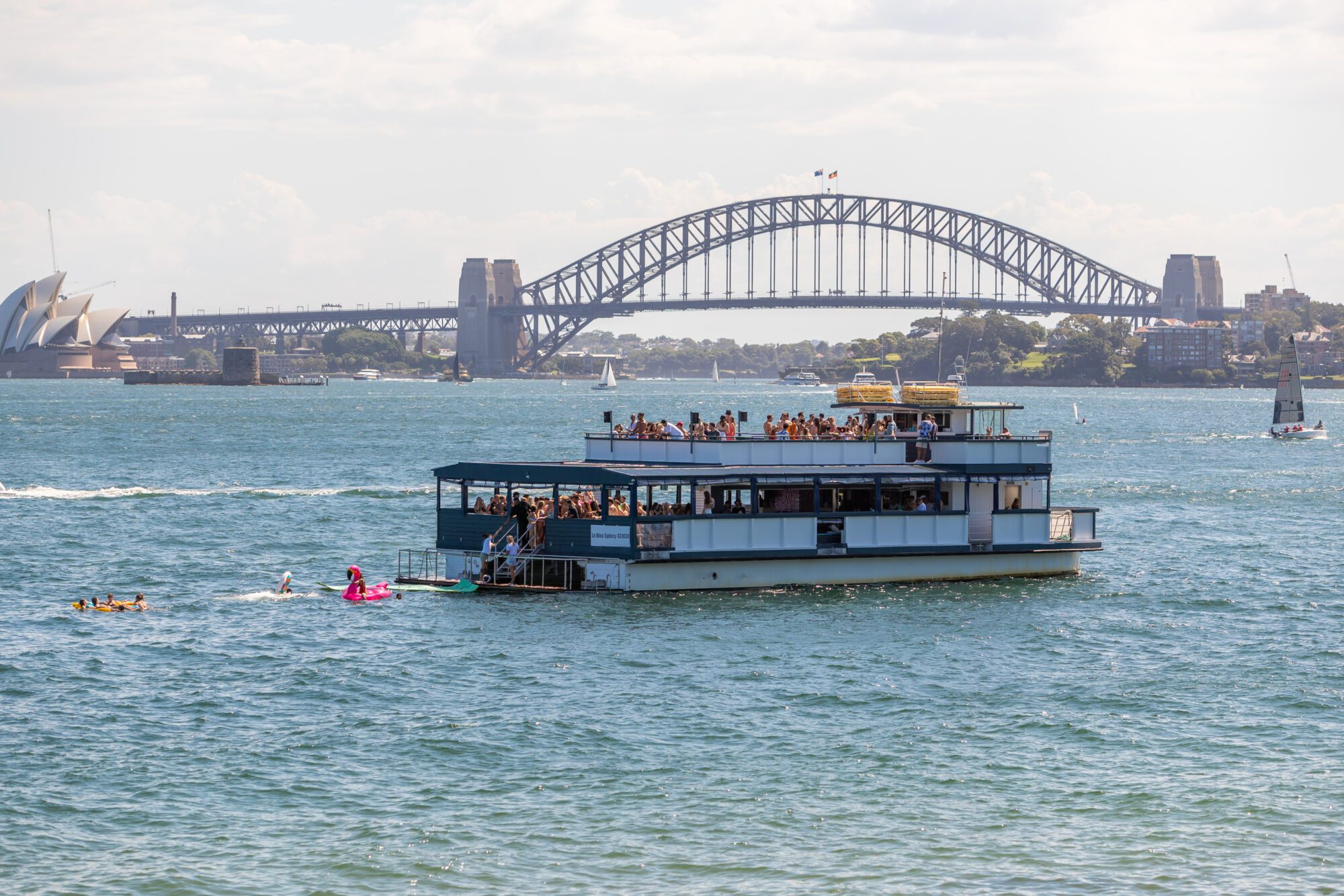 A boat with people on its upper deck floats on Sydney Harbour, with the Sydney Opera House and Sydney Harbour Bridge visible in the background under a partly cloudy sky. Swimmers are near the boat in the water.