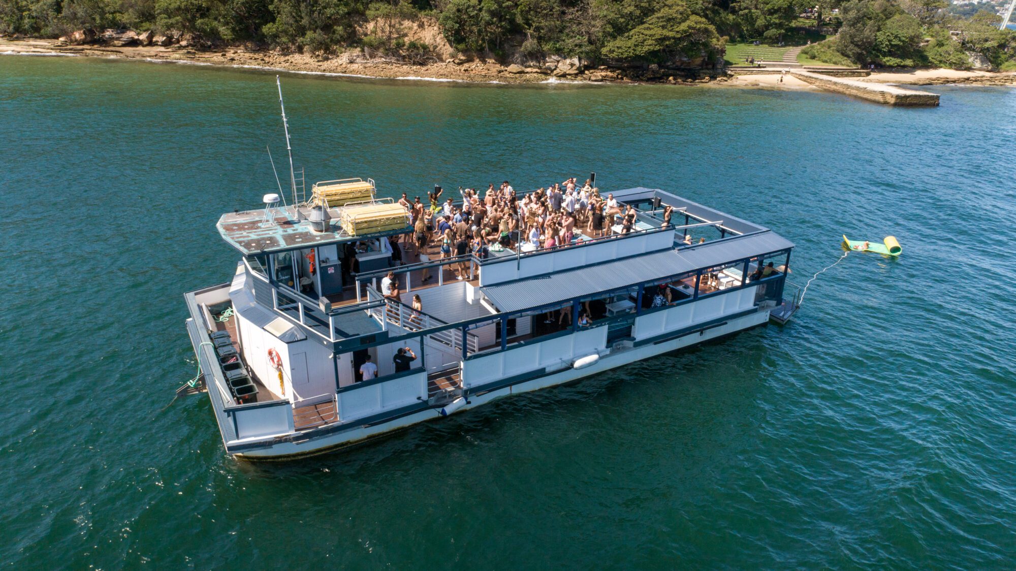 A large group of people gather on the upper deck of a boat in open water near a tree-lined shore, enjoying sunny weather and a party atmosphere.