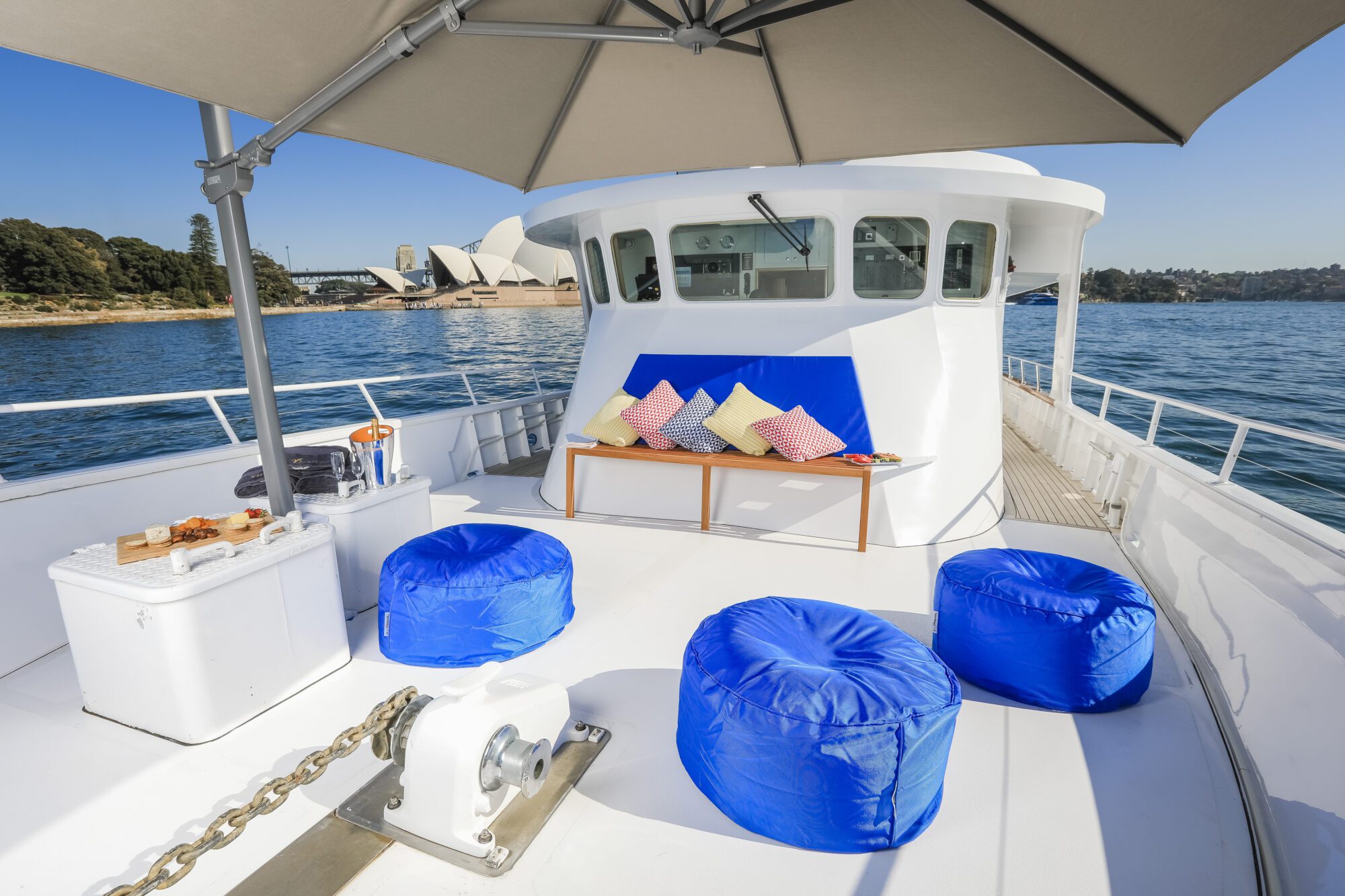 Sunny yacht deck with blue bean bags, white coolers, a shaded bench with colorful pillows, and the Sydney Opera House visible across the water.